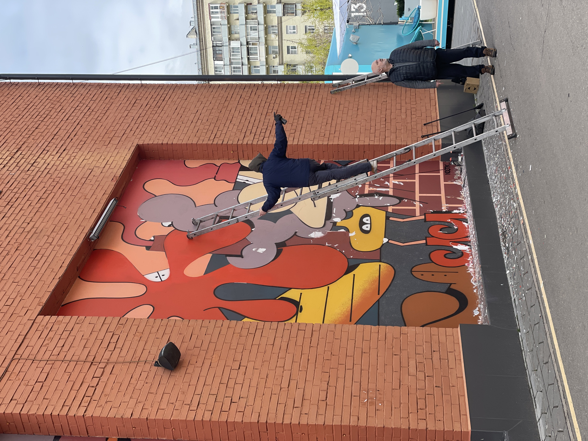 A street-level, medium-angle shot depicts two men working on a large mural in Khimki, Russia. The mural, set within a recessed brick wall, features an abstract, cartoonish design with dominant shades of red, brown, yellow, and grey. One man lies prone on an aluminum ladder, arm extended, presumably painting or touching up the mural. The second man stands on the ground near the base of another ladder, observing the other's work. A nearby building with multiple windows is visible in the background, along with a small blue structure with a ladder against it. The street appears to be recently cleaned, with some scattered debris near the mural.