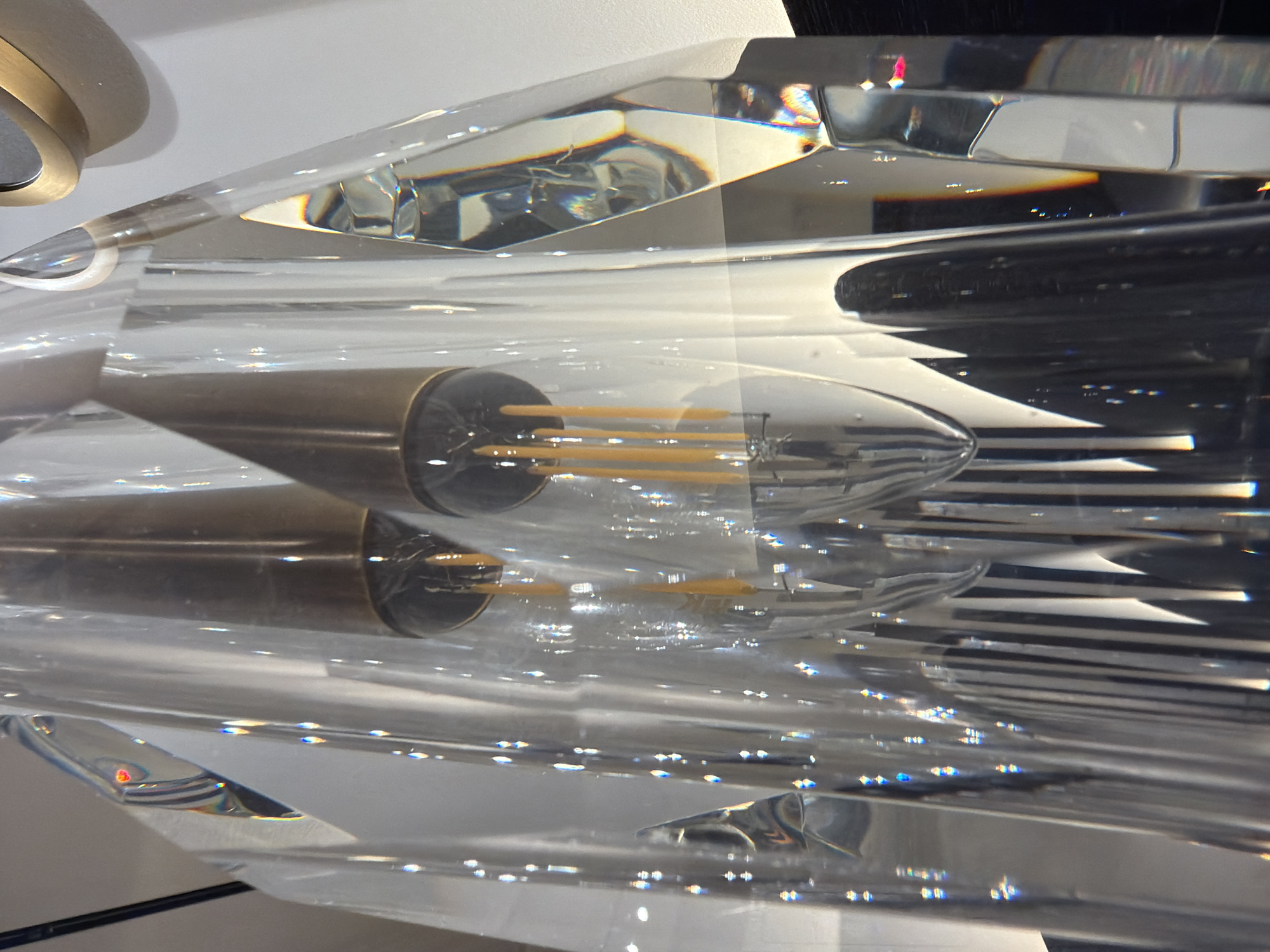 The image is a close-up, eye-level shot of a modern chandelier made of clear crystal and brass. The chandelier features multiple light bulbs with exposed filaments and brass caps, encased within faceted crystal prisms. The lighting in the image is dramatic, with bright points of light reflecting off the crystal surfaces, creating rainbow-like refractions. The background is a plain white wall. The chandelier appears to be hanging indoors, possibly in a showroom or a high-end residence, given its ornate and contemporary design. The exact location in the city of Unknown, Unknown is not discernible from the image itself.