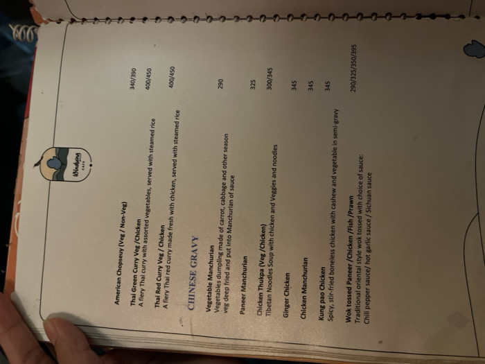 The image displays a slightly angled view of an open, spiral-bound menu from "Windbeng CAFE," presumably located in Dibrugarh, India. A hand is partially visible on the bottom left, holding the menu. The overall setting appears to be indoors, likely a restaurant or cafe, with soft lighting.
The menu features a variety of Asian cuisine, primarily Thai and Chinese dishes, with prices listed in a three-digit format, likely Indian Rupees. The logo for "Windbeng CAFE" is prominent on the left side, depicting a stylized fruit, possibly a blueberry or plum, with the cafe name written inside.
Visible text on the menu includes:
- **American Chopseuy (Veg/Non-Veg):** 340/390
- **Thai Green Curry Veg/Chicken:** A fiery Thai curry with assorted vegetables, served with steamed rice - 400/450
- **Thai Red Curry Veg/Chicken:** A fiery Thai red curry made fresh with chicken, served with steamed rice - 400/450
- **CHINESE GRAVY**
- **Vegetable Manchurian:** Vegetables dumpling made of carrot, cabbage and other season veg deep fried and put into Manchurian of sauce - 290
- **Paneer Manchurian:** 325
- **Chicken Thukpa (Veg/Chicken):** Tibetan Noodles Soup with chicken and Veggies and noodles - 300/345
- **Ginger Chicken:** 345
- **Chicken Manchurian:** 345
- **Kung pao Chicken:** Spicy, stir-fried boneless chicken with cashew and vegetable in semi-gravy - 345
- **Wok tossed Paneer/Chicken/Fish/Prawn:** Traditional oriental style wok tossed with choice of sauce: Chili pepper sauce/hot garlic sauce / Sichuan sauce - 290/325/350/395
The image captures a moment of menu browsing, offering a clear view of the food selections and their corresponding prices.