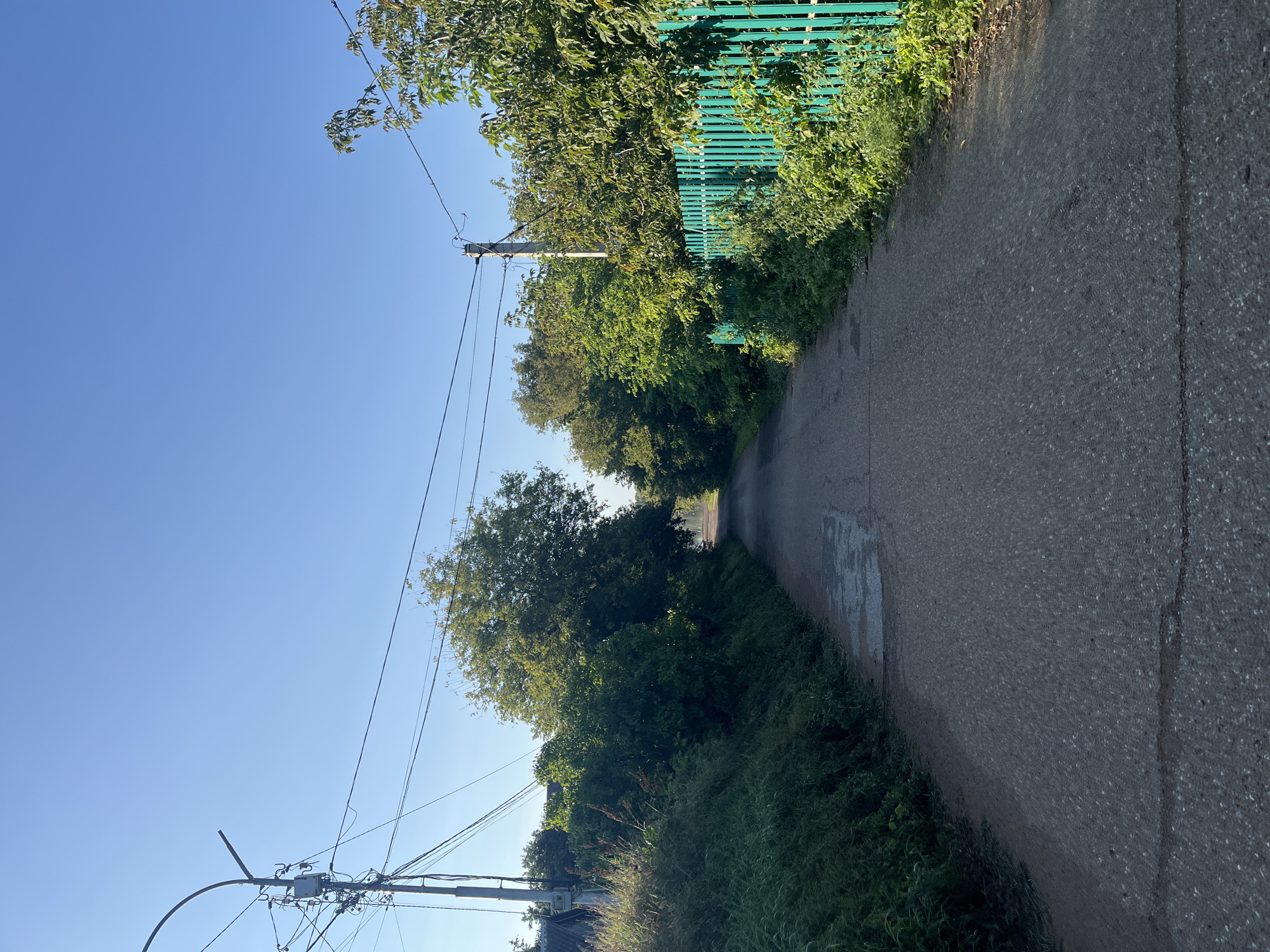 The image depicts a view down a narrow asphalt road, likely in a rural or suburban area. The road is flanked by lush green vegetation and trees on both sides. On the right side of the road, a bright turquoise-colored metal fence runs along the property line. A utility pole with numerous wires is visible on the left side of the road, and another utility pole stands on the right side, further down the road. The sky is a clear, vibrant blue, indicating good weather. The road surface shows signs of wear, with some patches of water or dampness visible. The overall impression is of a quiet, sunlit day in a green, somewhat unkempt, setting. There are no people or vehicles visible in the frame. This scene is consistent with a residential street in Blagoveshchensk, Russia, potentially a dacha settlement or a less developed urban area.