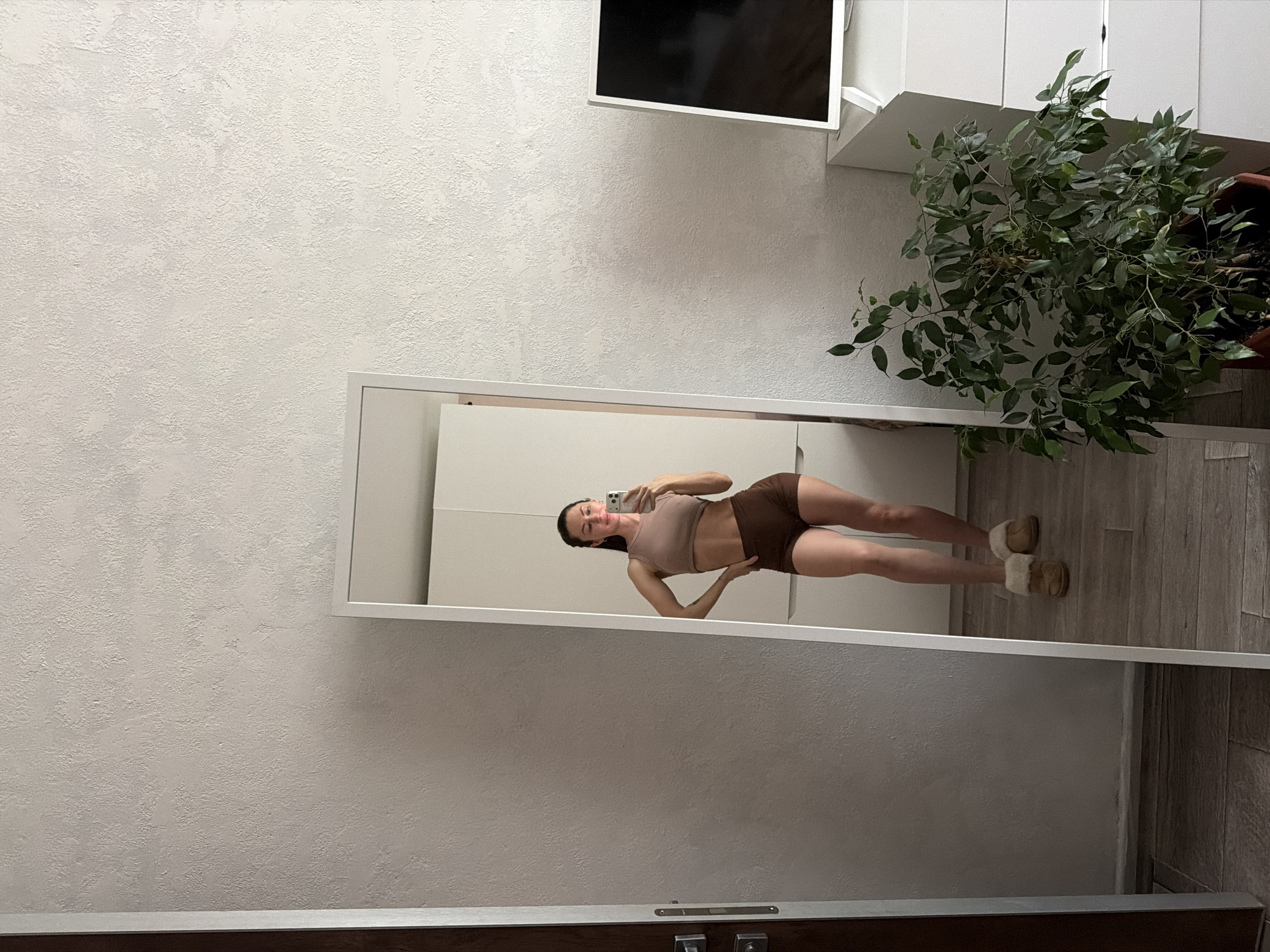 A young woman is taking a selfie in a full-length mirror. She is wearing a tan top and brown shorts with brown slippers. She is standing on a wooden floor with a white wall and white furniture in the background. A plant is placed to the right of the mirror. A TV is mounted on the wall in the upper center of the frame. The image was taken in Perm, Russia.