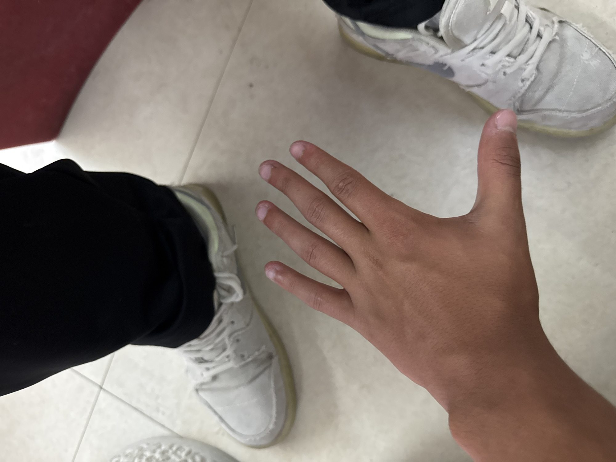 The image captures an overhead view of an indoor setting, focusing on a human hand in the foreground and two pairs of feet/shoes.
In the center foreground, a right hand is extended, palm facing slightly upwards. The skin tone is medium-brown, and the fingernails are short and unpolished. The knuckles and creases on the palm are visible.
In the upper right portion of the frame, the left foot of one individual is visible, wearing a light-colored, possibly off-white, athletic shoe with laces. The shoe appears somewhat worn. Below this, the lower part of a black pant leg is seen. In the lower left, the right foot of another individual is visible, also wearing a similar light-colored athletic shoe and a black pant leg. This shoe also shows signs of wear, particularly around the sole.
The individuals are standing on a light-colored, possibly off-white or beige, tiled floor with visible grout lines. A red object, possibly part of a wall or furniture, is visible in the upper left corner of the frame. There is no visible text in the image. The scene suggests a casual indoor environment, possibly in Yerevan, Armenia, as indicated by the location context.