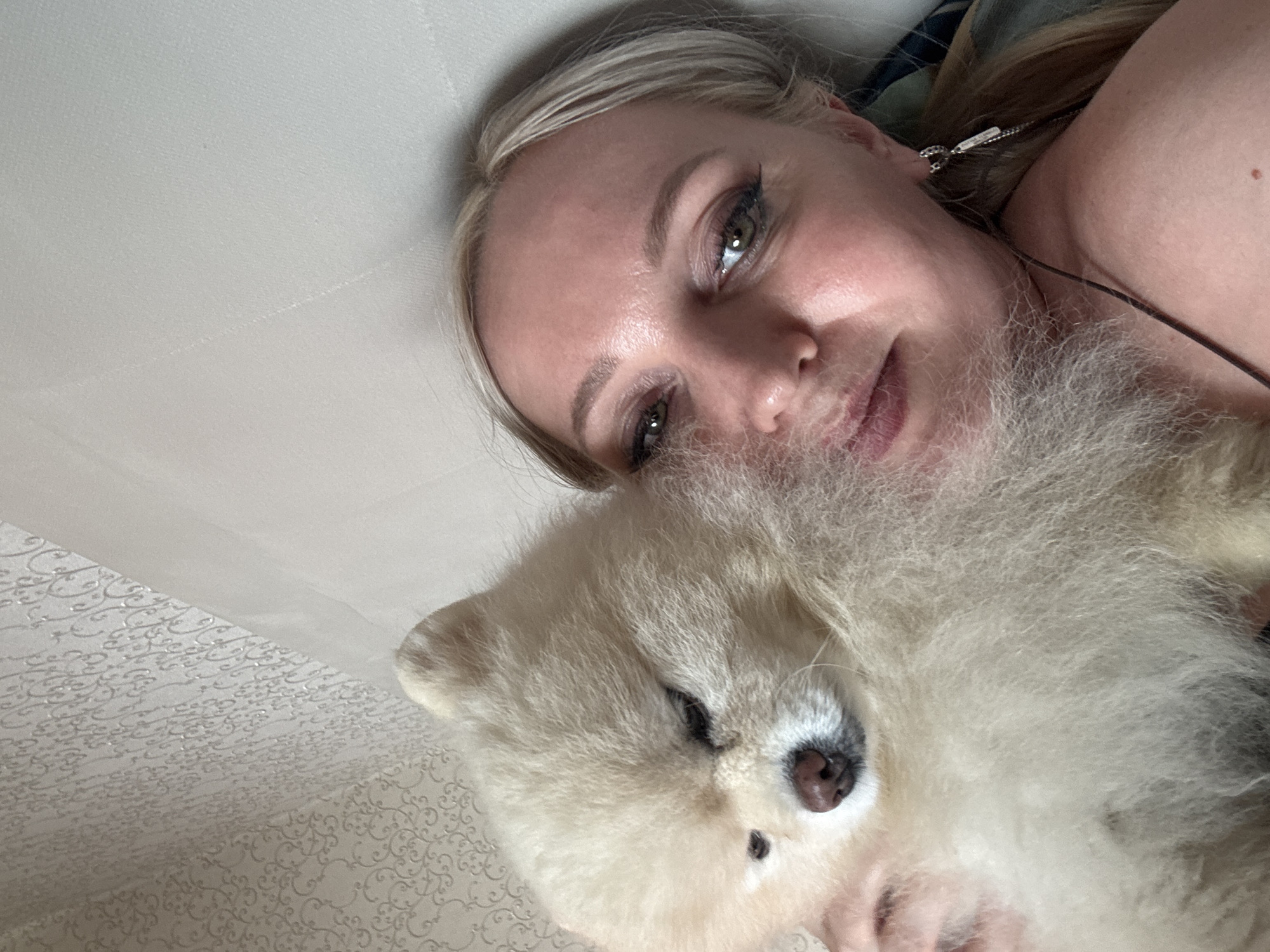 A blonde woman with green eyes and heavy eye makeup is lying down, embracing a light-colored Pomeranian dog. The dog has its eyes partially closed and its nose is touching the woman's face. The background is a textured wallpaper with a light pattern and a plain wall. The scene appears to be indoors, possibly a bedroom, in Komsomolsk-on-Amur, Russia, given the context provided.