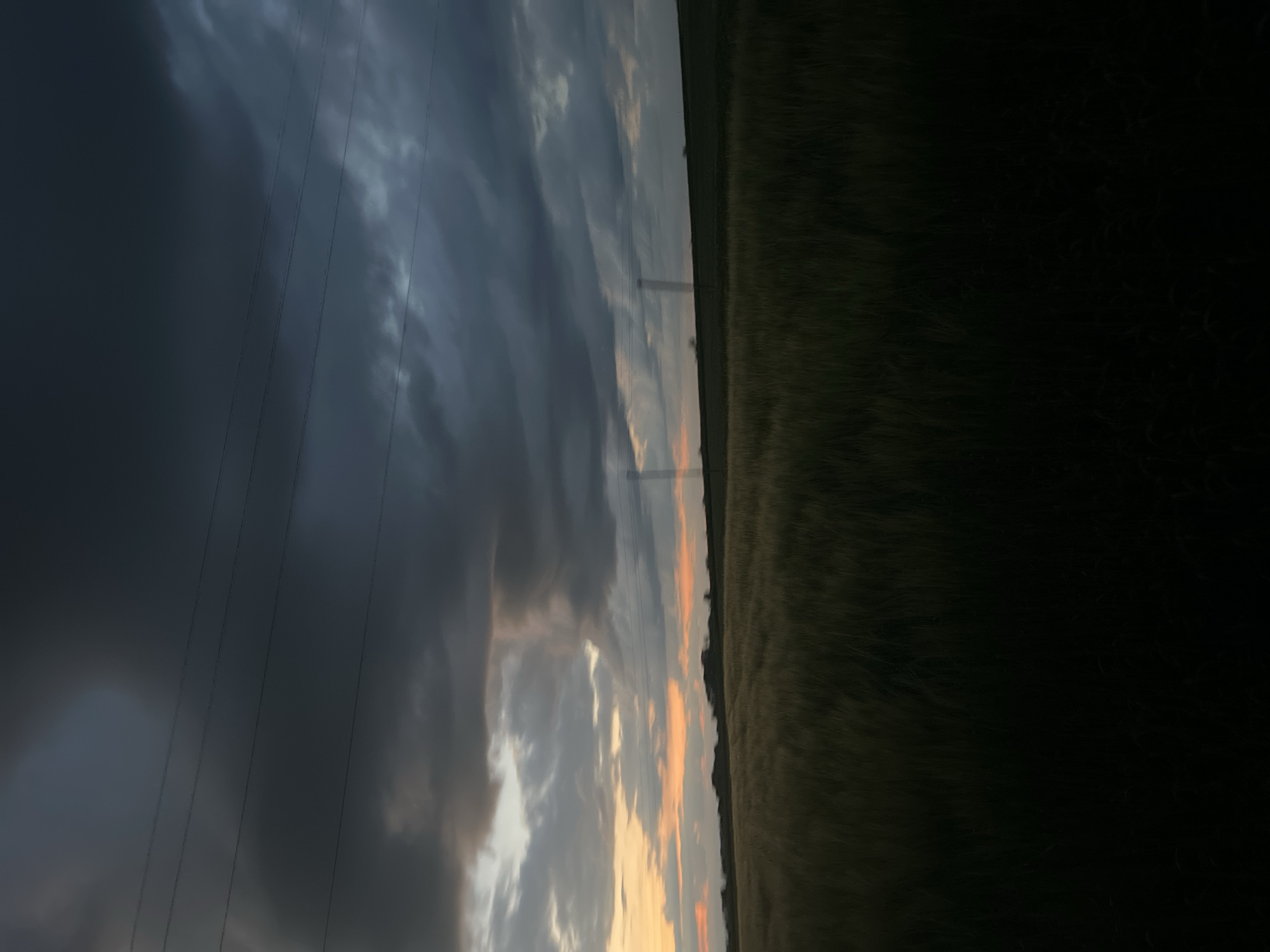 The image depicts a rural landscape under a dramatic, overcast sky. In the foreground, a dark field of tall grass or crops stretches across the lower half of the frame. The texture of the vegetation is visible, appearing dense and slightly wind-swept.
The middle ground features a slightly elevated horizon line. Several indistinct shapes, possibly trees or small buildings, dot the landscape. A few faint structures that appear to be utility poles or pylons are also visible on the horizon, with power lines stretching across the sky.
The sky dominates the upper portion of the image. Heavy, dark clouds create a brooding atmosphere, with streaks of orange and yellow light breaking through near the horizon, suggesting a sunset or sunrise. The overall lighting is dim, with the dark clouds and foreground field contributing to a somber mood. The location is identified as Zarudyntsi, Ukraine. No people or specific actions are depicted.