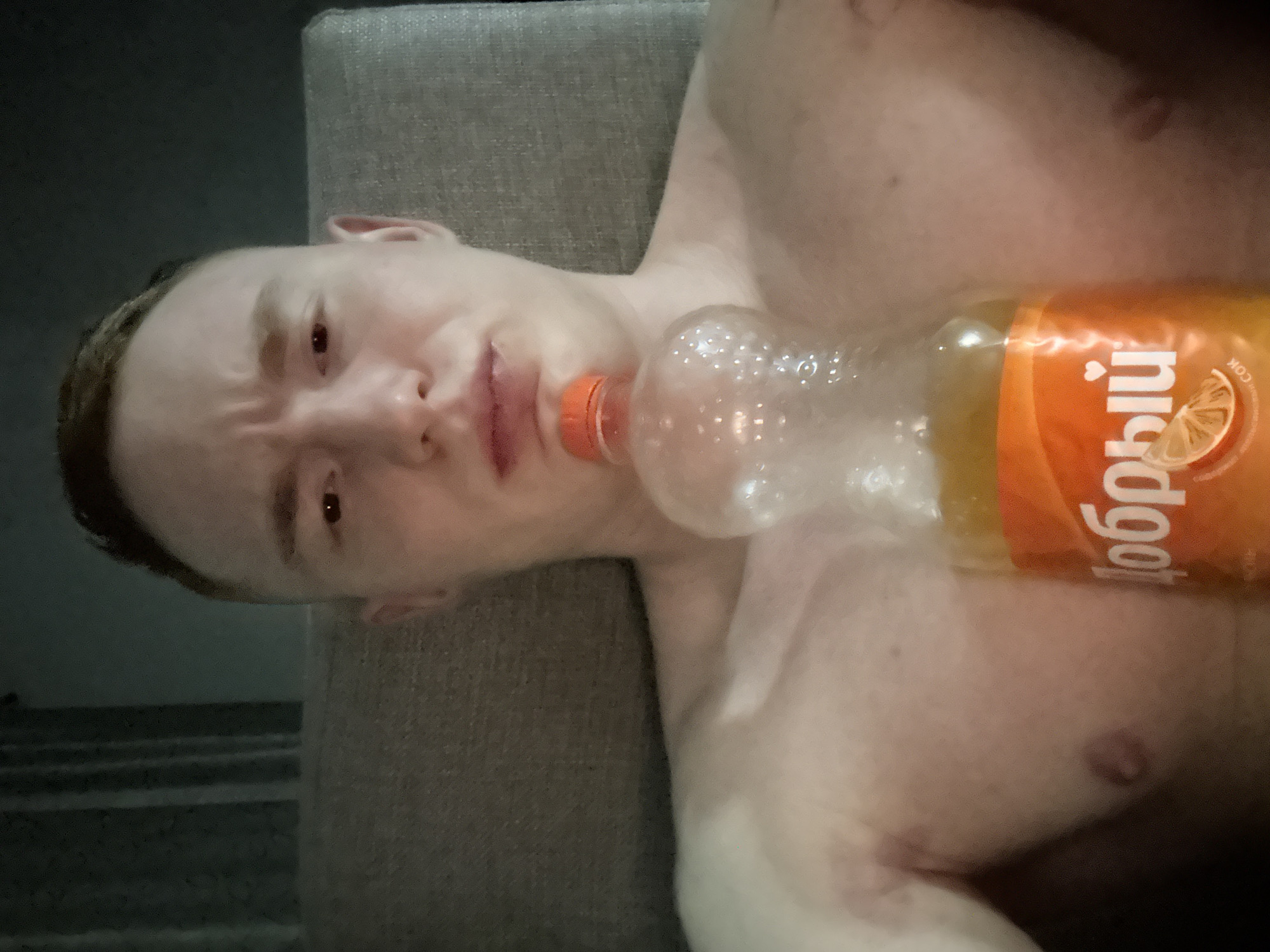 A man is lying on a grey sofa in a dimly lit room. His bare chest is visible. He has fair skin, short brown hair, and a furrowed brow. An almost empty bottle of "Dobry" orange juice is positioned diagonally across his chest. The location is presumed to be Chaikovsky, Russia, based on the bottle's brand and Russian text.