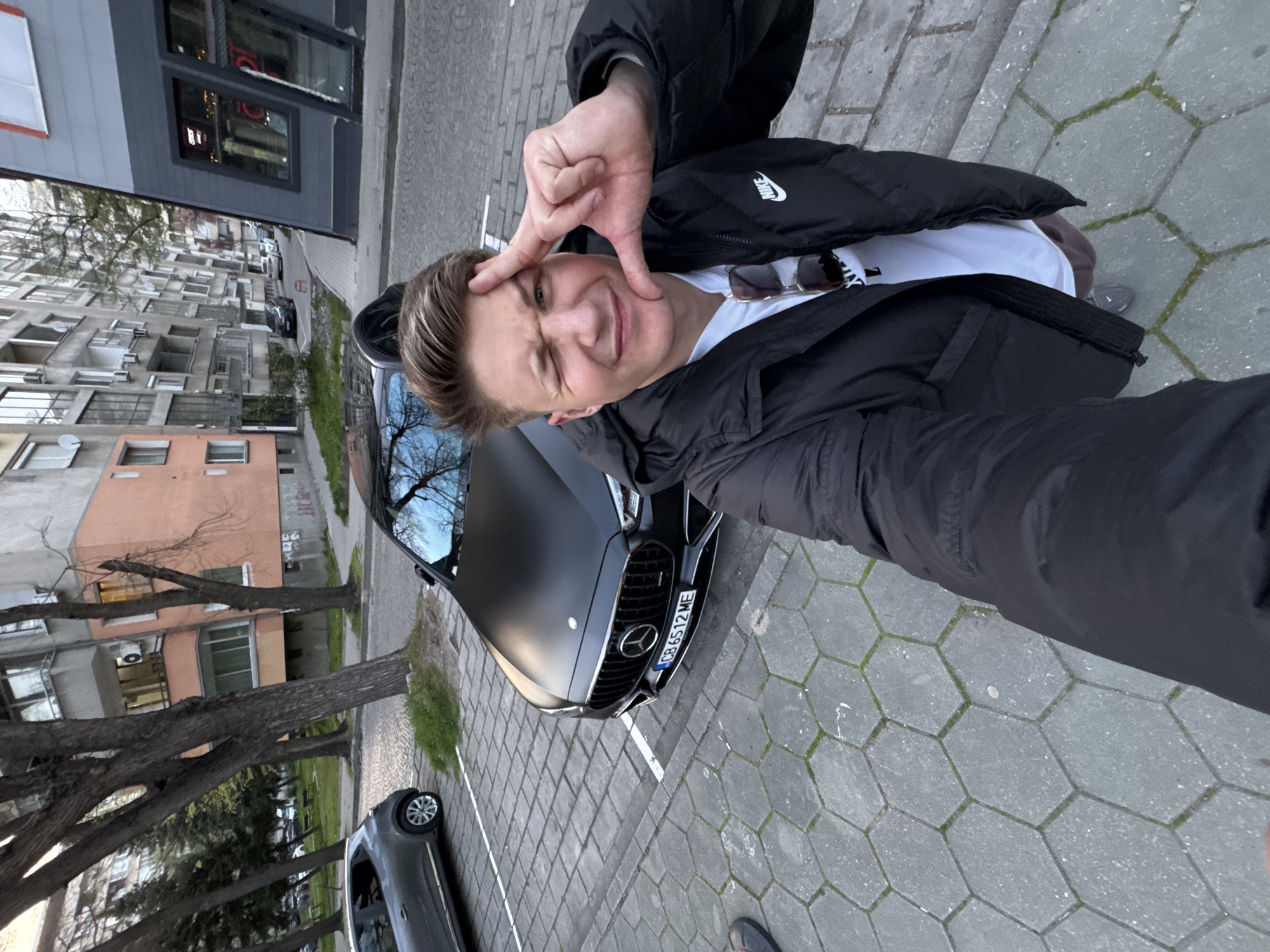 A man stands in front of a matte black Mercedes-Benz car, posing for a selfie. He wears a black Nike jacket over a white t-shirt and makes a square gesture with his hand near his face. Sunglasses rest on the t-shirt. The car has a license plate that reads "CB 6512 ME." The street is paved with grey hexagonal stones, and there is another car parked to the left, further down the street. In the background, there is a building with windows, suggesting an urban setting. Based on available information, the scene is likely located in Plovdiv, Bulgaria.