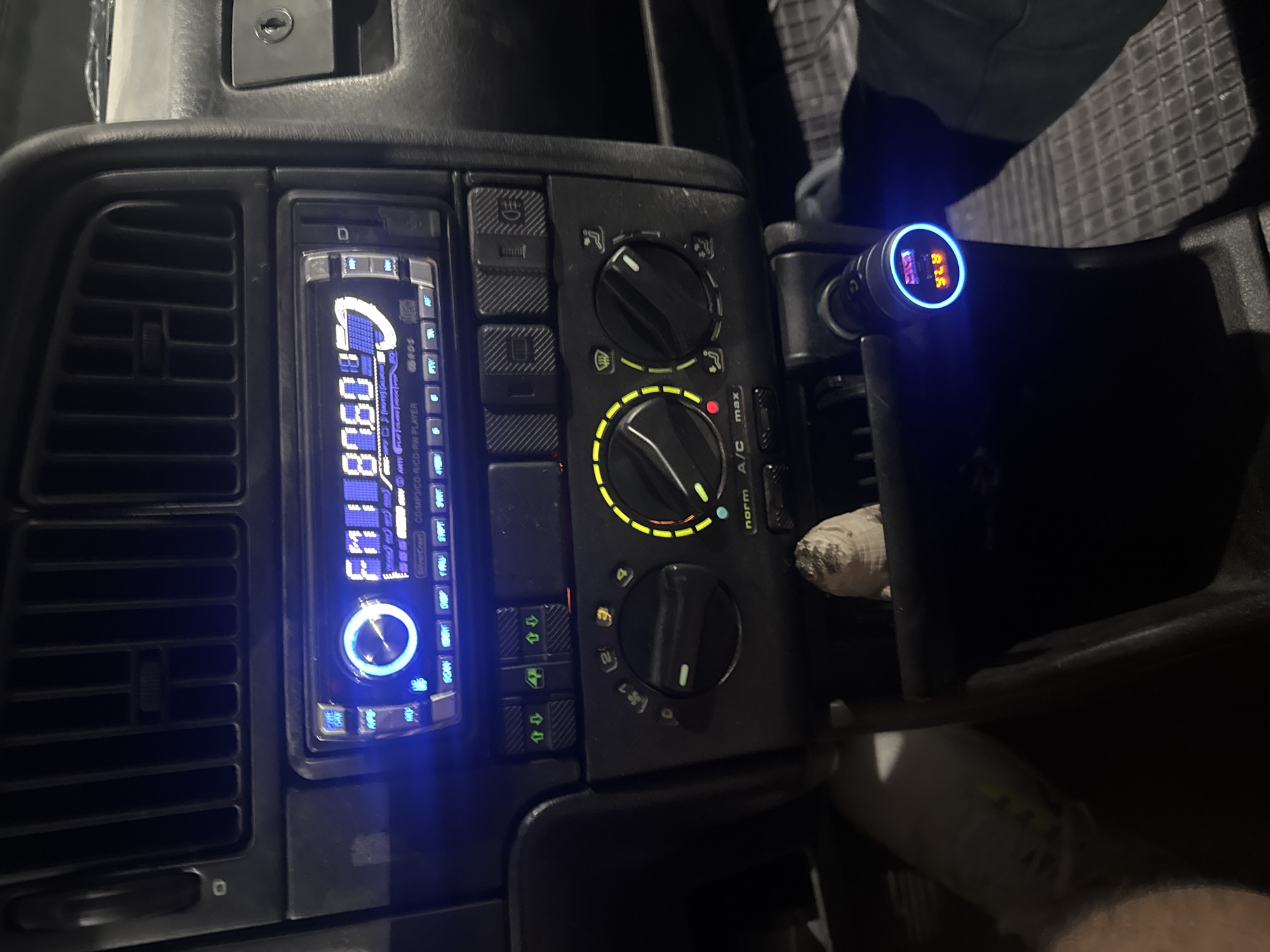This image shows the dashboard of a car, likely taken at night or in low light. The dashboard is black with a digital car radio tuned to 87.60 FM on the left. Next to the radio are the car's climate control knobs and buttons. A device with a blue illuminated display showing "12.5" is plugged into what appears to be the cigarette lighter or auxiliary power outlet. It is difficult to determine the exact car model. Based on the image content, there's no clear information to determine the exact location in Plovdiv, Bulgaria.