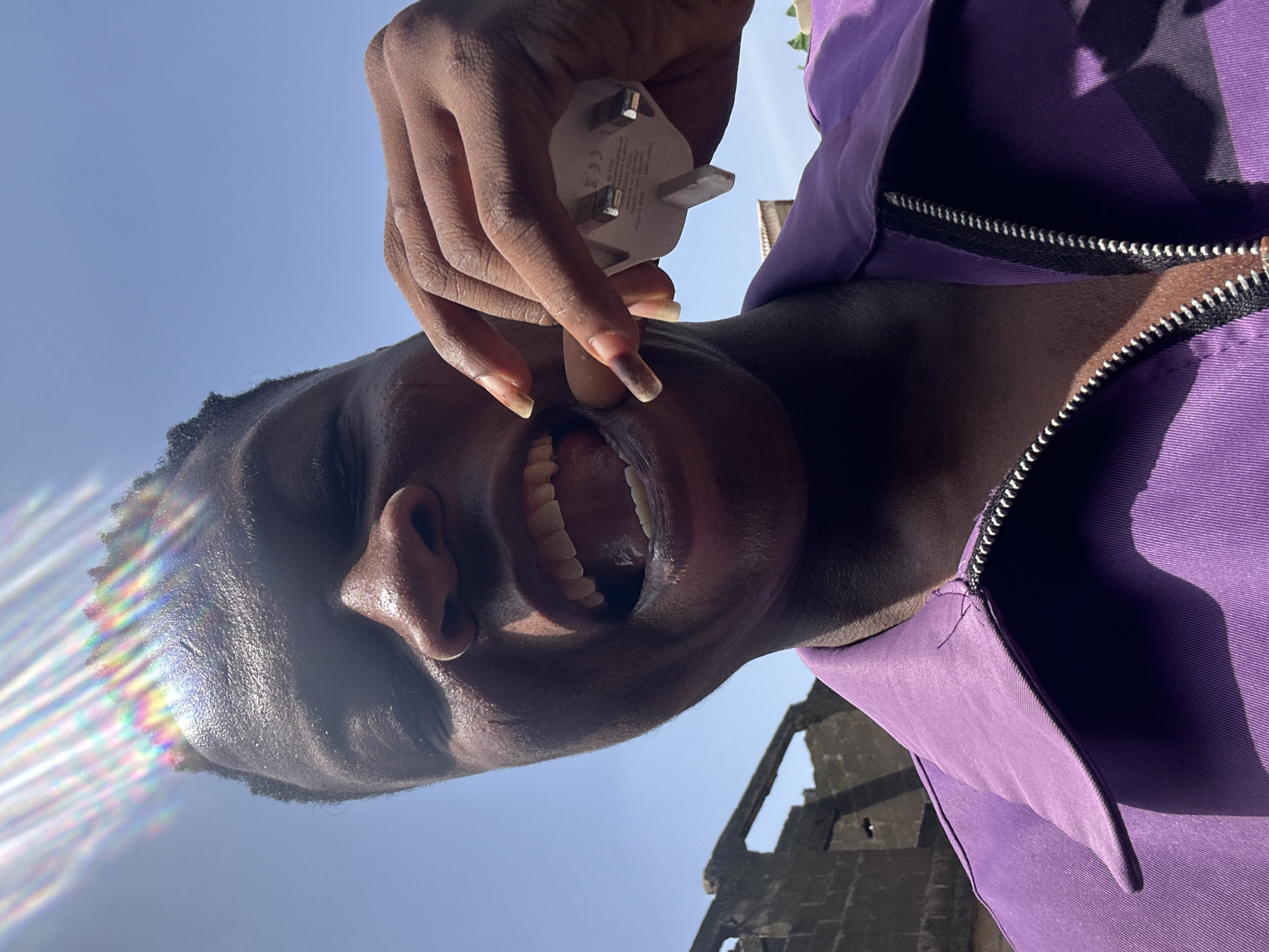 A person is holding a white UK electrical plug near their open mouth. The person is wearing a purple jacket with a black zipper. The background shows a clear blue sky and a partially visible ruin-like structure. The location is Ikorodu, Nigeria.