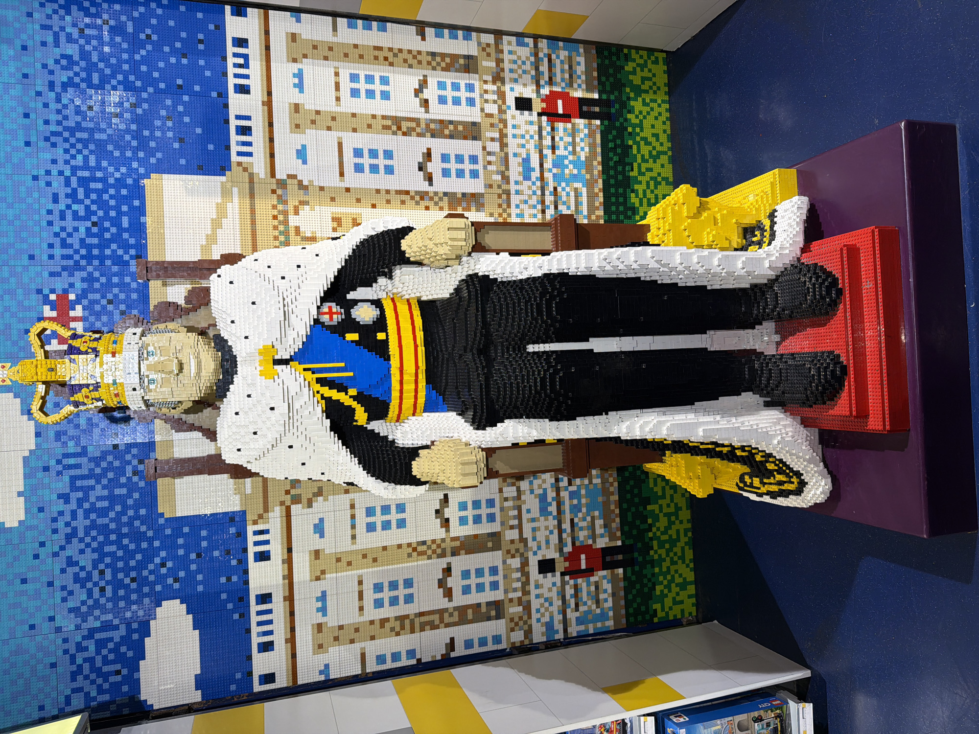 A Lego sculpture of King Charles III in coronation regalia stands in front of a Lego representation of Buckingham Palace in London, United Kingdom. The sculpture is on a raised purple and red platform. The King is wearing a white and black robe with a golden crown. The background shows Buckingham Palace in front of a blue sky. The scene is indoors.
