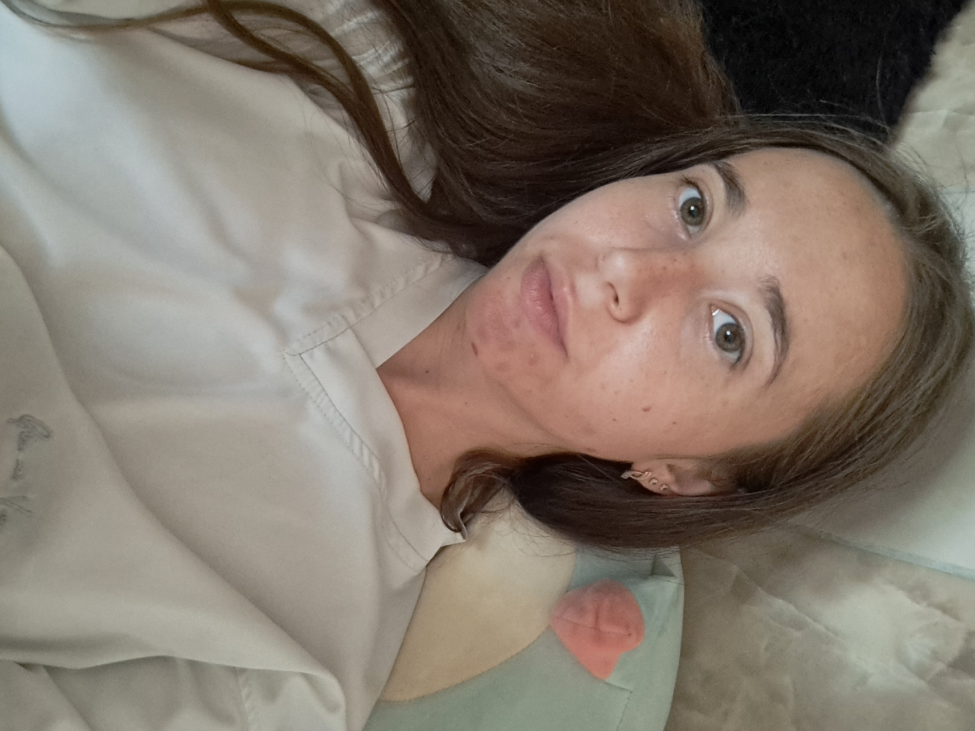 A close-up, overhead selfie of a young woman with long, brown hair lying on her back. She is wearing a light-colored, V-neck top. Her face is turned towards the camera, with her eyes open and looking forward. There are a few blemishes on her chin and cheek. Her hair is falling to her left side. Behind her, a light-colored padded surface is visible, along with a light green cushion with a pink heart-shaped object on it. The lighting appears to be natural, coming from above. The scene is indoors. There is no discernible location in the city of Vypasne, Ukraine, within the provided image.
