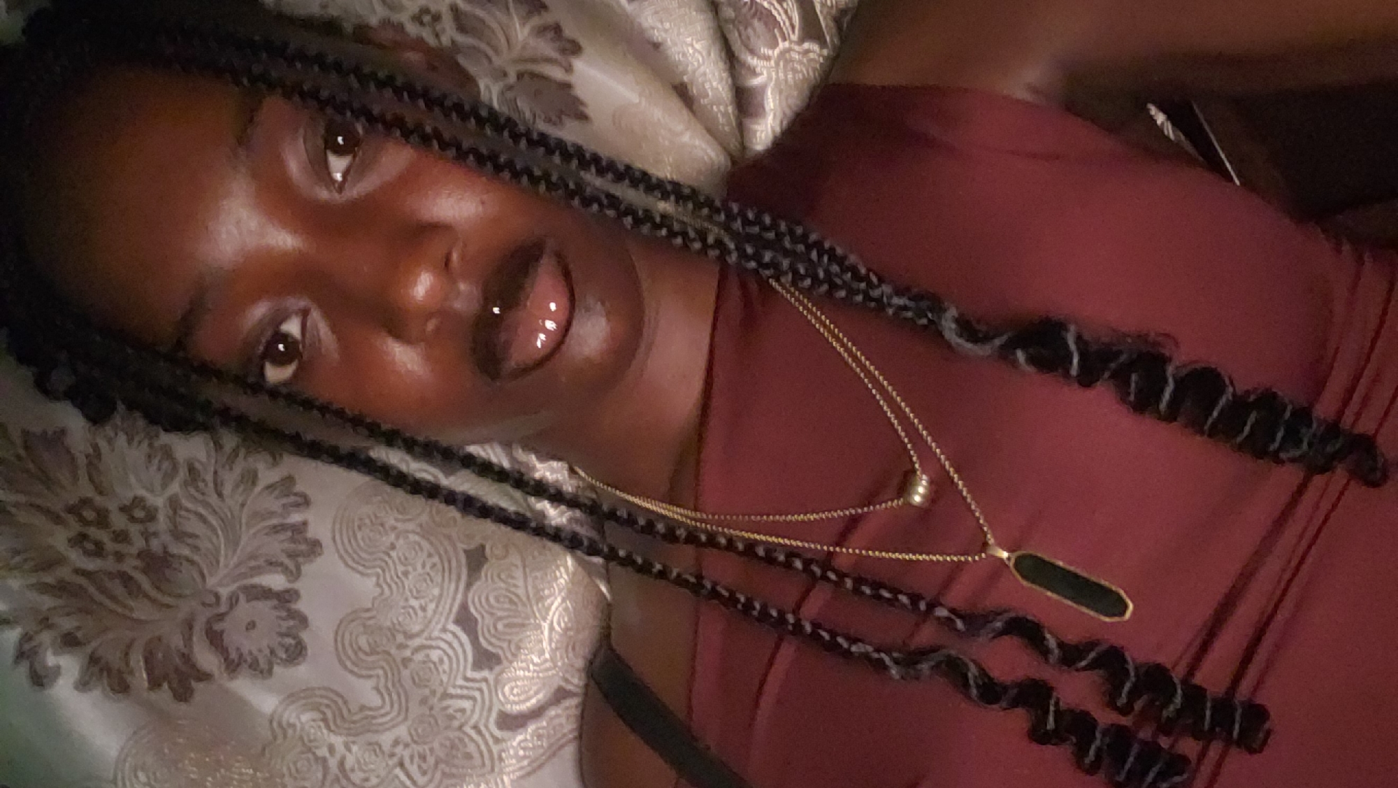 A close-up selfie shows a young Black woman with braided hair and a nose ring. She is wearing a maroon top and two necklaces, one with a gold pendant. The background is a textured, light-colored fabric. The image appears to have been taken indoors. There is no identifiable information to suggest the location is Rido, Nigeria.