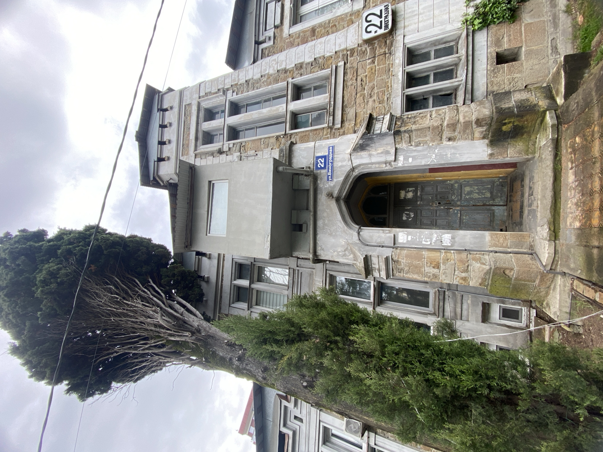 The image depicts the exterior of a building located at 22 Vinogradnaya Street in Sochi, Russia. The building appears to be old with stone masonry and a grey painted facade. The entrance features an arched doorway with a recessed door. The building has multiple windows with a rectangular design, and address plaques with the number "22" are visible on the building's exterior. A tree with both green foliage and bare branches obscures a portion of the lower floors. The sky is overcast.