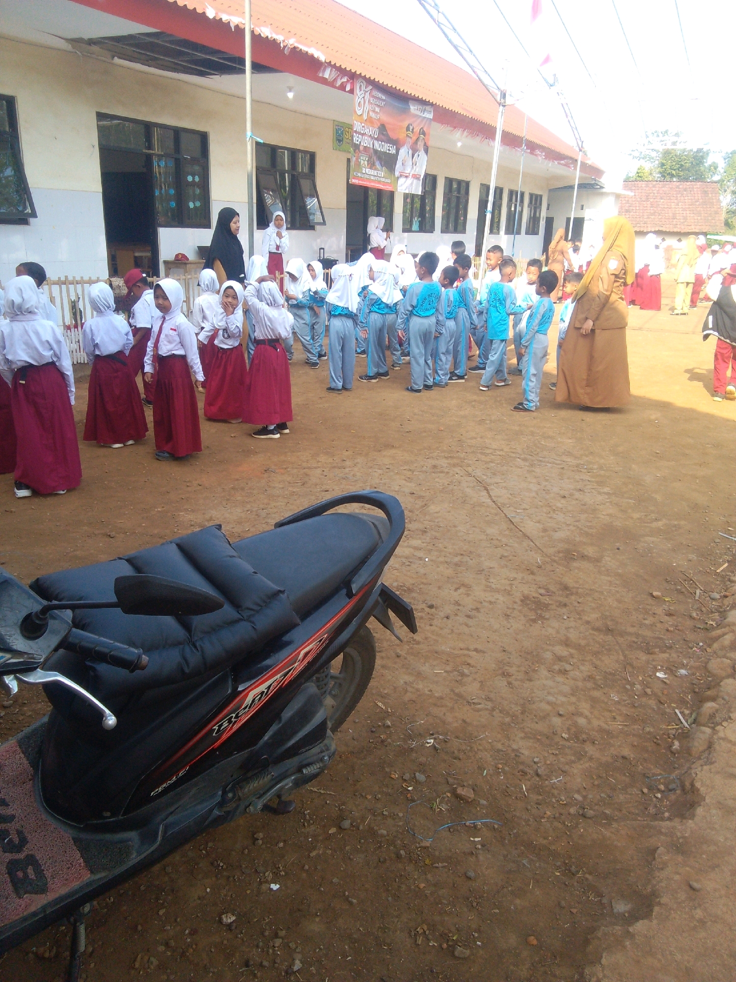 The image depicts a schoolyard scene in the city of Betek, Indonesia, on a sunny day. A group of young students, mostly girls in red skirts and white tops with hijabs, are gathered in front of a school building with a red and white awning. Some are standing in line, while others appear to be interacting. Two adult women are also present; one, wearing a black hijab and dress, stands near the school entrance, and another in a tan hijab and long brown dress stands further back. Several boys in light blue uniforms are also present, some facing the camera. A motorcycle with a black seat is parked in the foreground, partially obscuring the ground, which is dirt and gravel. In the background, the school building has multiple windows, and a banner with text is visible above. The overall atmosphere suggests a school event or assembly.