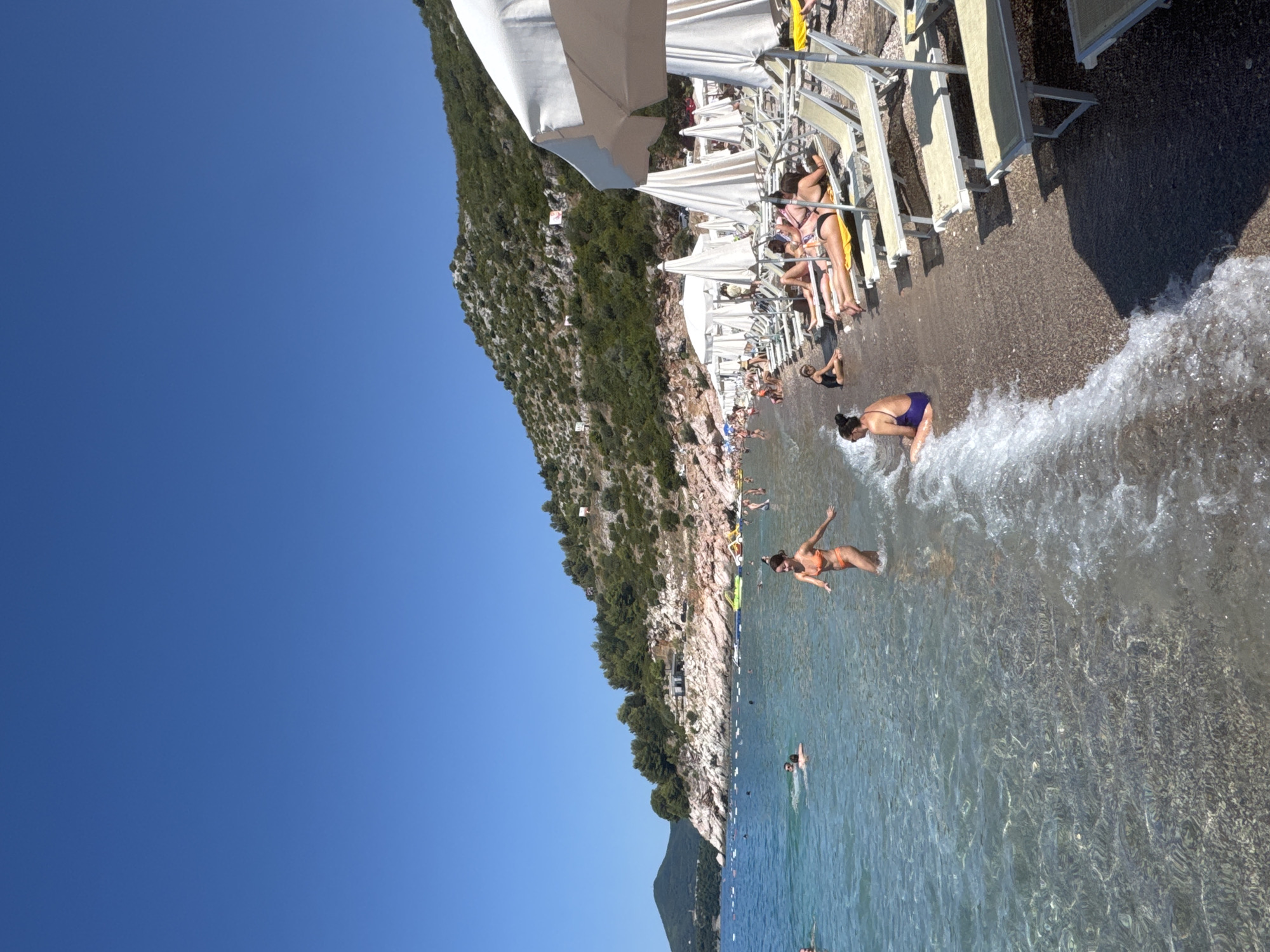The image depicts a sunny day at Kamenovo beach in Kamenovo, Montenegro. A clear blue sky stretches above a sloping, green, and rocky hillside that forms the backdrop of the beach. The beach itself is covered in pebbles.
In the foreground, the clear, shallow waters of the Adriatic Sea gently lap the shore. Several people are visible in the water, swimming and wading. One woman in an orange bikini is walking in the shallow water towards the left of the frame. Further out in the water, two men are swimming.
The beach is lined with rows of empty and occupied lounge chairs, many covered by large white umbrellas. People are seen sunbathing on the lounge chairs. A woman in a purple bikini is sitting on the shore to the left, facing away from the camera.
In the middle ground, the hillside is covered in dense green vegetation and interspersed with rocky outcrops. Some small buildings or structures are visible on the hillside, along with what appear to be flags or signs. The overall scene is one of a busy summer day at a popular coastal resort.