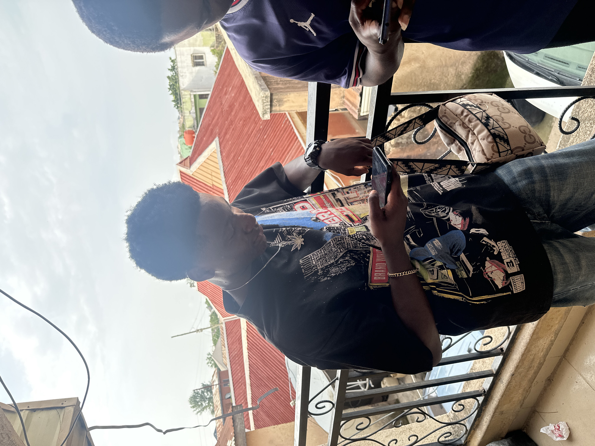 Two young Black men are standing on a balcony overlooking a residential area. The man on the left is wearing a black t-shirt with a graphic print and blue jeans. He is looking down at his phone, which he is holding in his left hand. He has a silver chain around his neck and a gold bracelet on his left wrist. A beige crossbody bag with a GG pattern is slung over his shoulder. The man on the right is wearing a dark blue t-shirt with a white Jumpman logo. He is also looking down at his phone. The balcony has a black metal railing with decorative scrollwork. In the background, there are buildings with red tiled roofs. The sky is overcast. The overall scene depicts a casual outdoor setting.