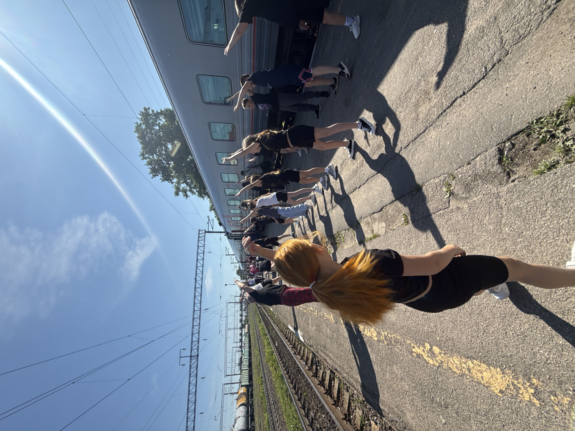 The image depicts an outdoor scene at a train station in Kanash, Russia, under clear, bright skies. A group of approximately ten to fifteen individuals, primarily young adults or teenagers, are lined up along a concrete train platform. They are engaged in a synchronized activity, standing with their backs mostly to the camera, extending their arms forward and slightly upwards in a uniform pose. Most are dressed in casual, dark-colored athletic wear, including t-shirts, shorts, and leggings. The person closest to the camera has long, reddish-blonde hair tied in a ponytail and is wearing a black top and black shorts.
To the left of the group, a light-colored passenger train is stopped parallel to the platform. The train features multiple windows along its side. Further left, beyond the platform, multiple railway tracks with gravel ballast extend into the distance, accompanied by overhead power lines supported by metal gantries against the sky. The sky is bright blue with a few wispy clouds, and a strong sun flare is visible in the upper left, indicating a sunny day. Long, distinct shadows of the people are cast across the platform, suggesting either early morning or late afternoon. The concrete platform shows some cracks and wear. No legible text is visible on the train or platform.