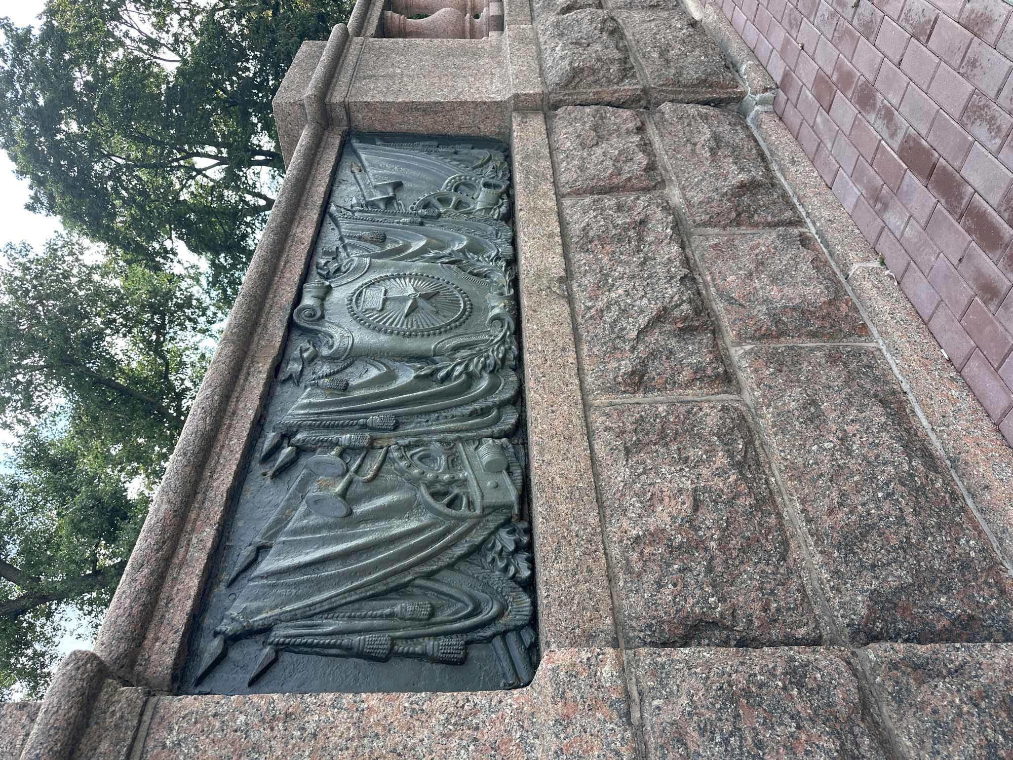 The image shows a close-up of a bronze relief on a granite wall in Minsk, Belarus. The relief depicts a frieze of military symbols, including flags, trumpets, and a coat of arms with a star. The wall is constructed from large, rough-hewn granite blocks. To the right of the wall, a red brick pathway is visible, with some wet patches indicating recent rain. Above the wall, lush green trees with dappled sunlight are seen against a bright sky. The relief is detailed and appears to be well-preserved, suggesting it is a historical or commemorative monument.