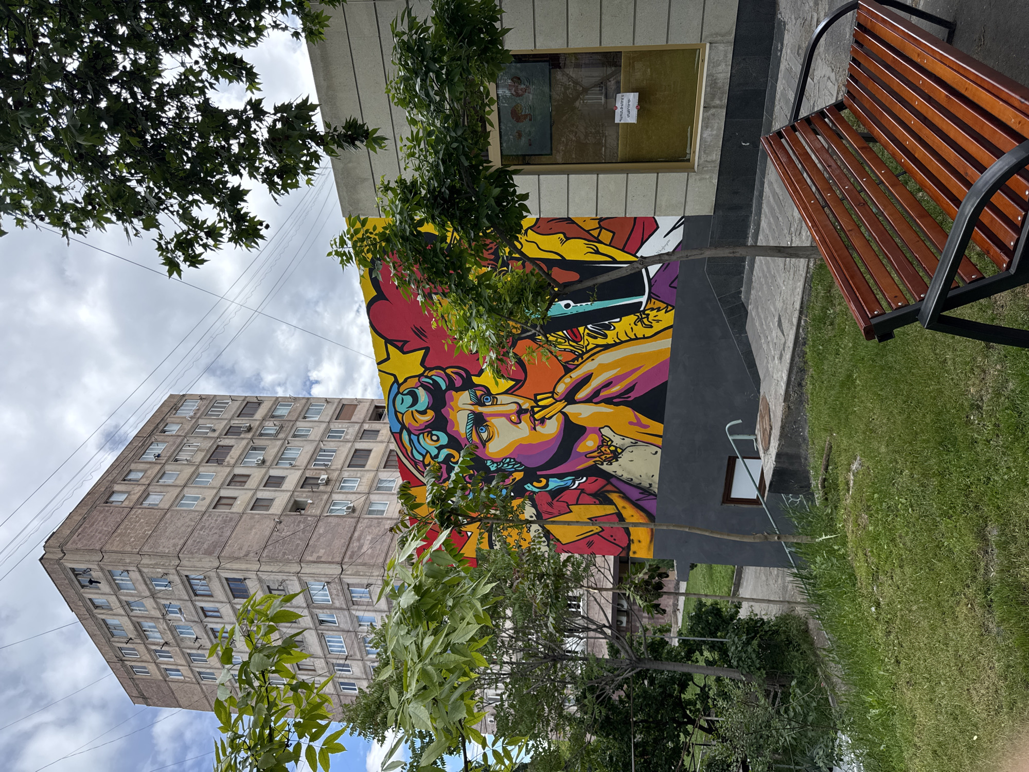 The image captures a vibrant mural adorning a building in Yerevan, Armenia. The mural, rich in color, depicts a person smoking and is positioned on the side of a building next to a window. A tall, Soviet-era apartment building stands in the background. Trees partially obscure the view of the mural and the apartment building. To the right, a wooden bench with a black metal frame sits on a patch of green grass. The sky is cloudy with blue patches visible. Overhead power lines can be seen stretching toward the apartment building.
