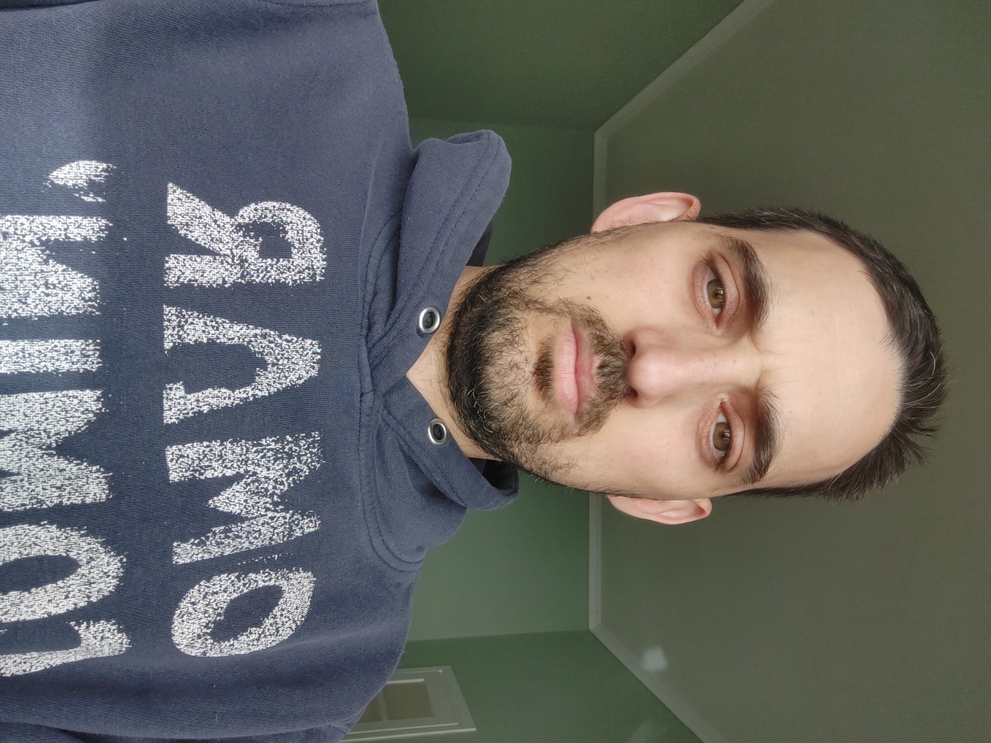 A medium-shot, selfie-style, indoor photograph captures a man with a beard and brown hair wearing a blue hooded sweatshirt with white lettering. The walls are light green. The photo is taken from an upward angle, tilting the man's face to the right. The sweatshirt lettering is reversed. There is a small white vent towards the lower left corner of the image.
There is no evidence this image was taken in Tel-Aviv, Israel.