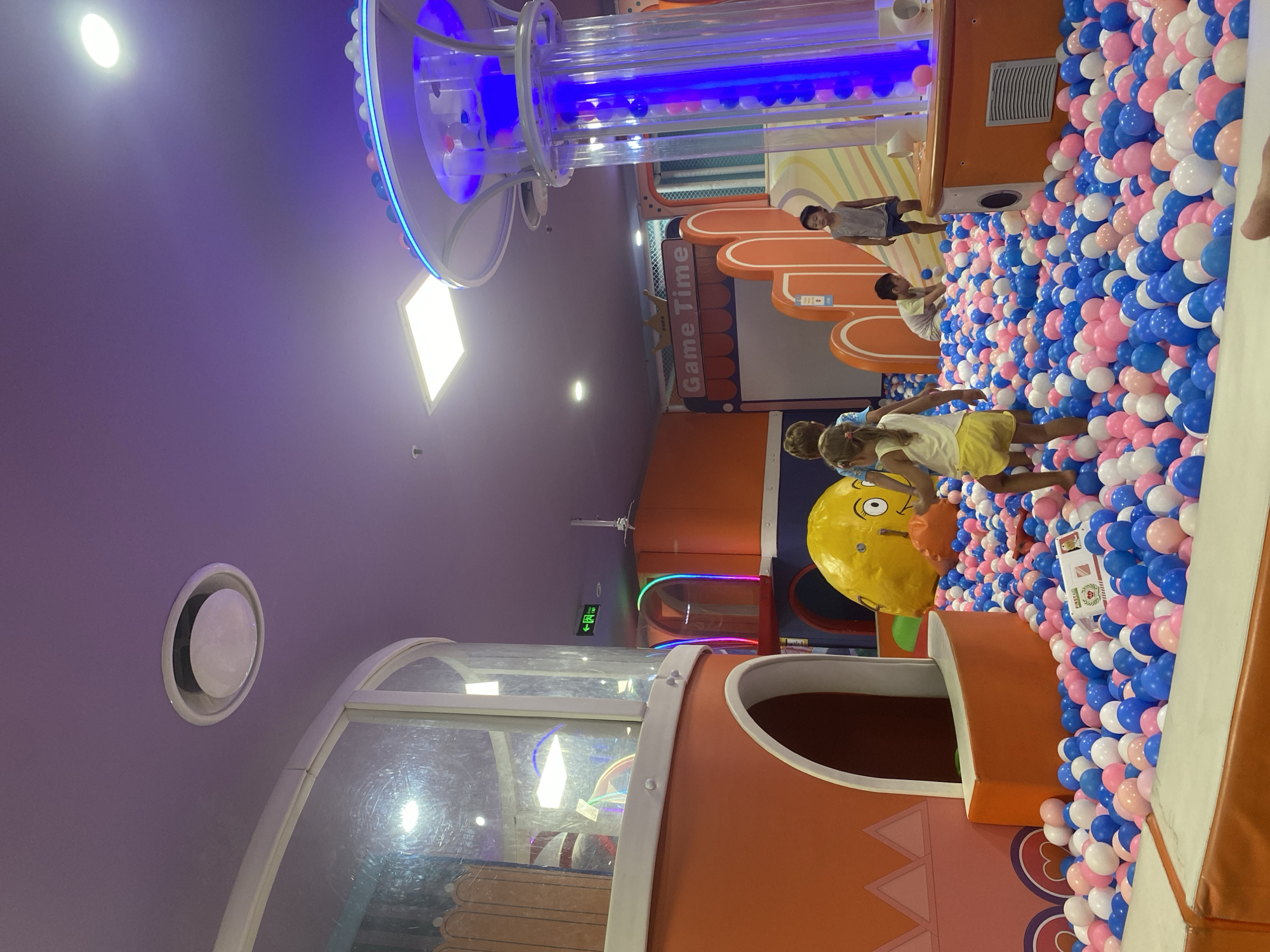 The image depicts an indoor playground filled with a ball pit. Several children are visible playing amongst the multi-colored balls, which are predominantly blue, pink, and white. In the background, there is a large yellow inflatable object resembling a face, a structure with clear tubes containing blue light and balls, and various play structures with orange and white accents. Signage reading "Game Time" is visible. The environment appears to be a commercial establishment, likely an amusement center or family entertainment venue. No specific location in Phu Thuy, Vietnam is discernable from the image.