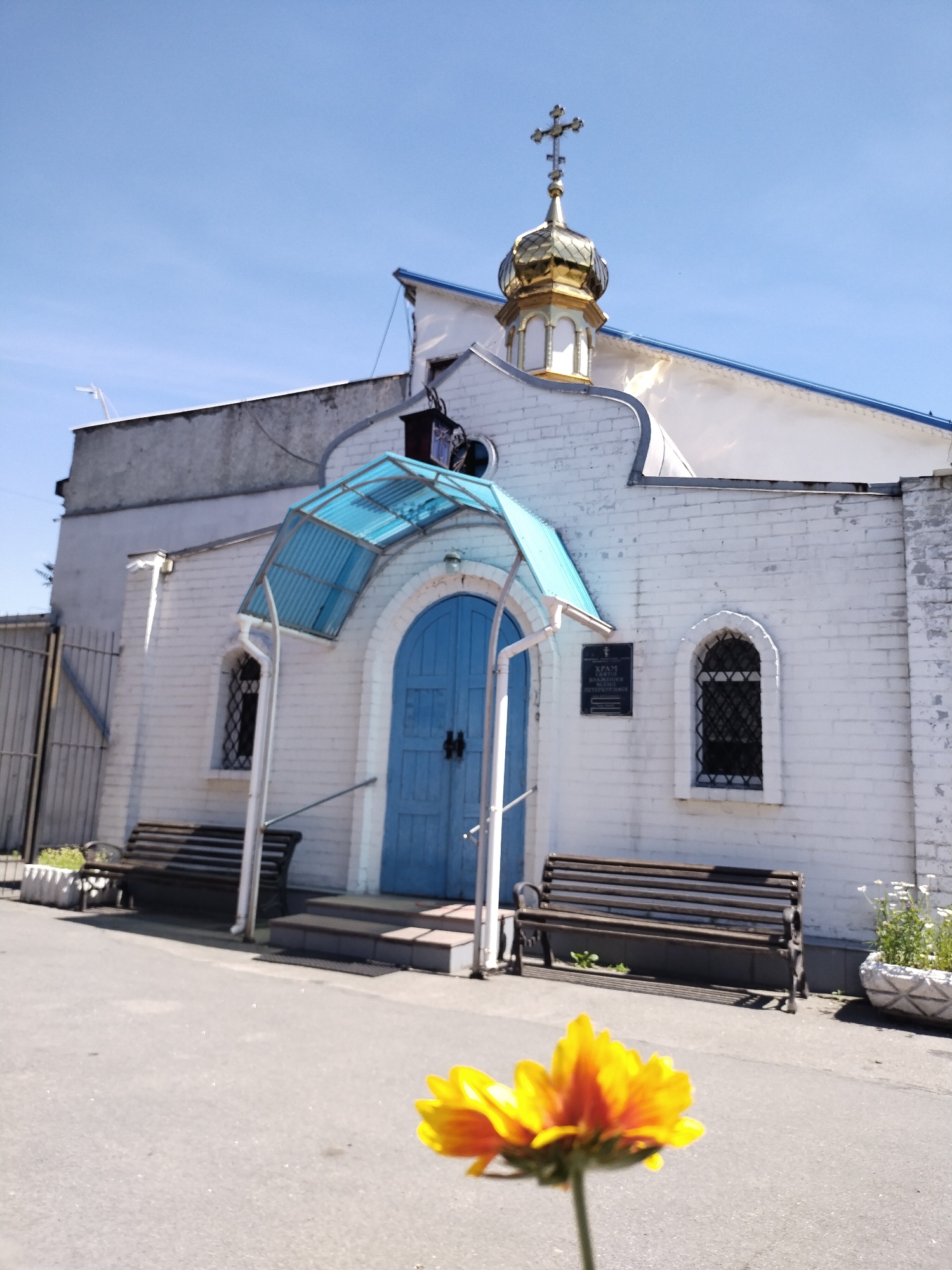 The image shows the entrance to a small, white brick church with a golden dome topped by a cross, located in the city of Horishni Plavni, Ukraine. The entrance features a blue door under a blue, corrugated plastic awning. Two wooden benches are situated to the left and right of the entrance. In the foreground, a single yellow and orange flower is in sharp focus, obscuring part of the view of the church grounds. The sky is clear and blue, and the scene appears to be set in daylight.