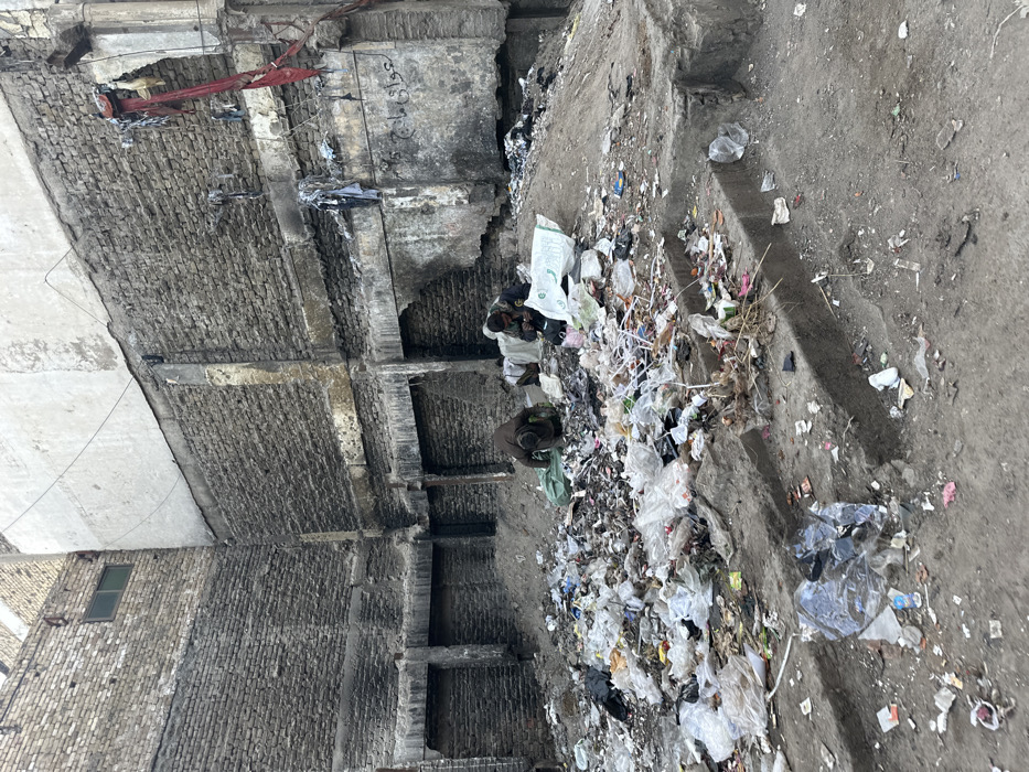 The image portrays a severely degraded urban area within Quetta City Tehsil, Pakistan. The ground in the mid-ground and foreground is covered by a substantial accumulation of mixed refuse, including numerous plastic bags, paper, and general debris, forming a large, irregular mound. Two adult males are present within this refuse heap. The individual on the left is stooped over, facing away from the viewer, seemingly sifting through the waste, with a green bag nearby. The individual on the right is crouched or seated, also appearing to sort items from the refuse, with a white bag positioned beside him.
The background consists of three dilapidated walls forming an interior corner. These walls are constructed predominantly of weathered, exposed brick, displaying extensive signs of decay, charring, and structural compromise, particularly evident in their lower sections where mortar is eroded and bricks are damaged. A section of the upper right wall features light-colored plaster over brickwork, contrasting with the exposed brick. A small, horizontally oriented, dark-framed window is visible high on the left brick wall. Inscriptions, identifiable as Arabic or Urdu script, are present on the lower part of the right brick wall. Red fabric and other indeterminate debris hang from the upper-right section of the same wall. Concrete steps, partially obscured by ground debris, are visible in the lower right foreground. The scene is illuminated by ambient daylight.