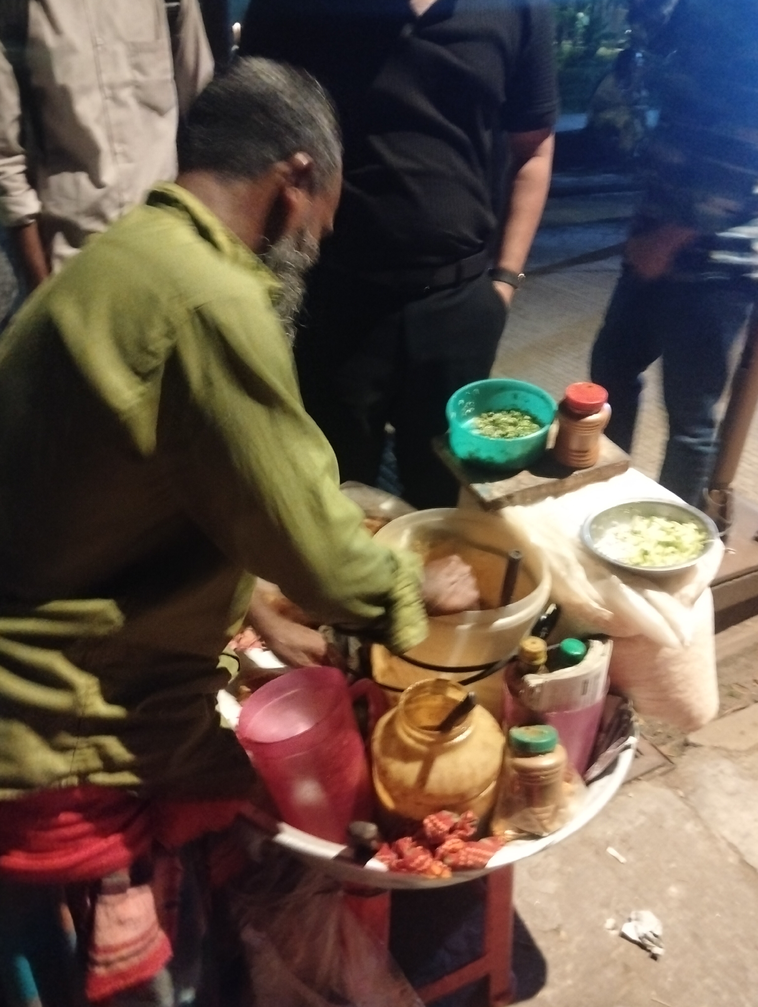 A street vendor with a beard is preparing food from a cart in Dhaka, Bangladesh. The cart contains various ingredients and condiments, including diced vegetables, spices, and sauces. The vendor is wearing a green shirt and a red and patterned cloth around his waist. There are three other people nearby, two of whom are standing near the cart and one further back. The setting appears to be outdoors at night on a paved surface.