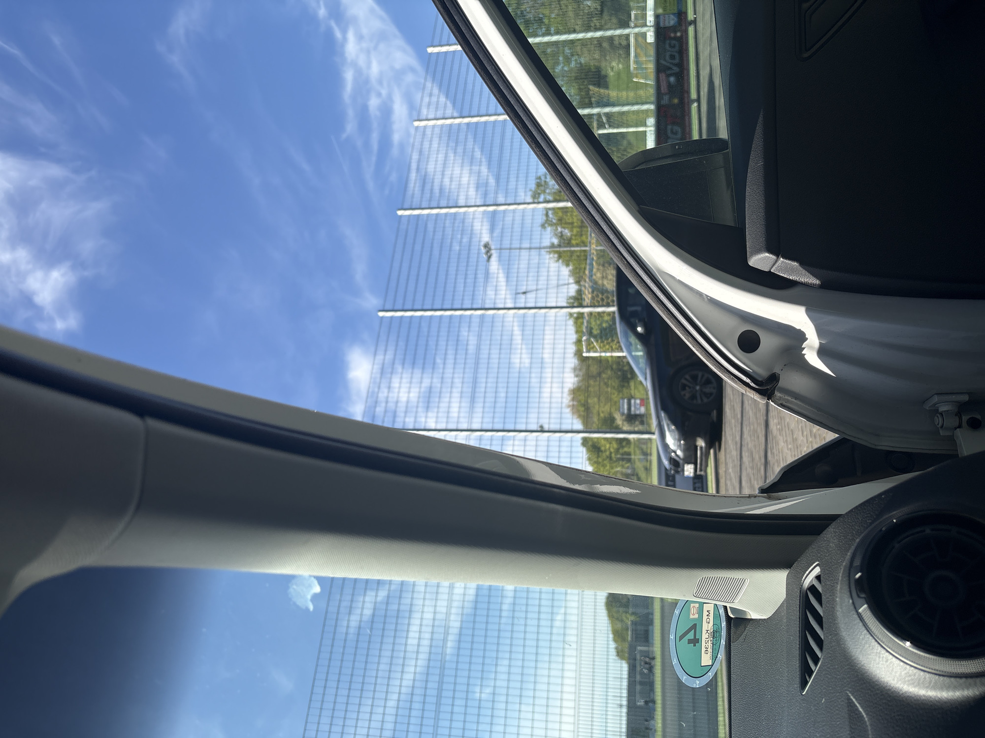 The image, taken from inside a white car, shows the exterior of the vehicle through the open car door. A black car is visible outside. A large, tall metal grid-like structure stretches across the background, likely a sound barrier. The sky is blue with scattered clouds. A green circular sticker with white lettering is visible on the lower left portion of the windshield. Based on the vehicle license plate sticker, the location is Wassenberg, Germany.