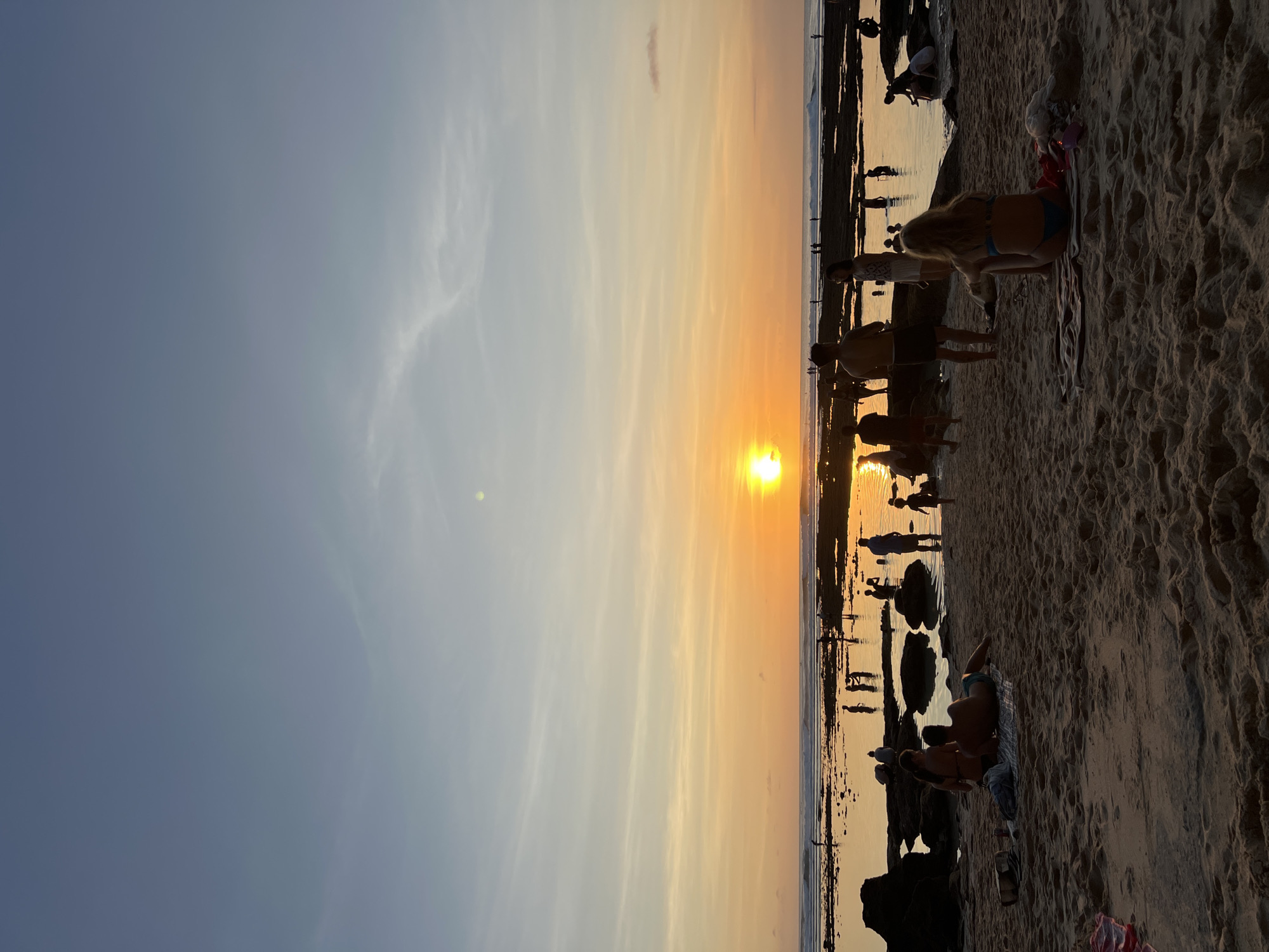 The image shows a beach scene at sunset. The sun is setting over the ocean, casting an orange glow on the sky. Silhouettes of people dot the shoreline. In the foreground, several individuals are relaxing on the sand. The location is Unknown, Unknown.