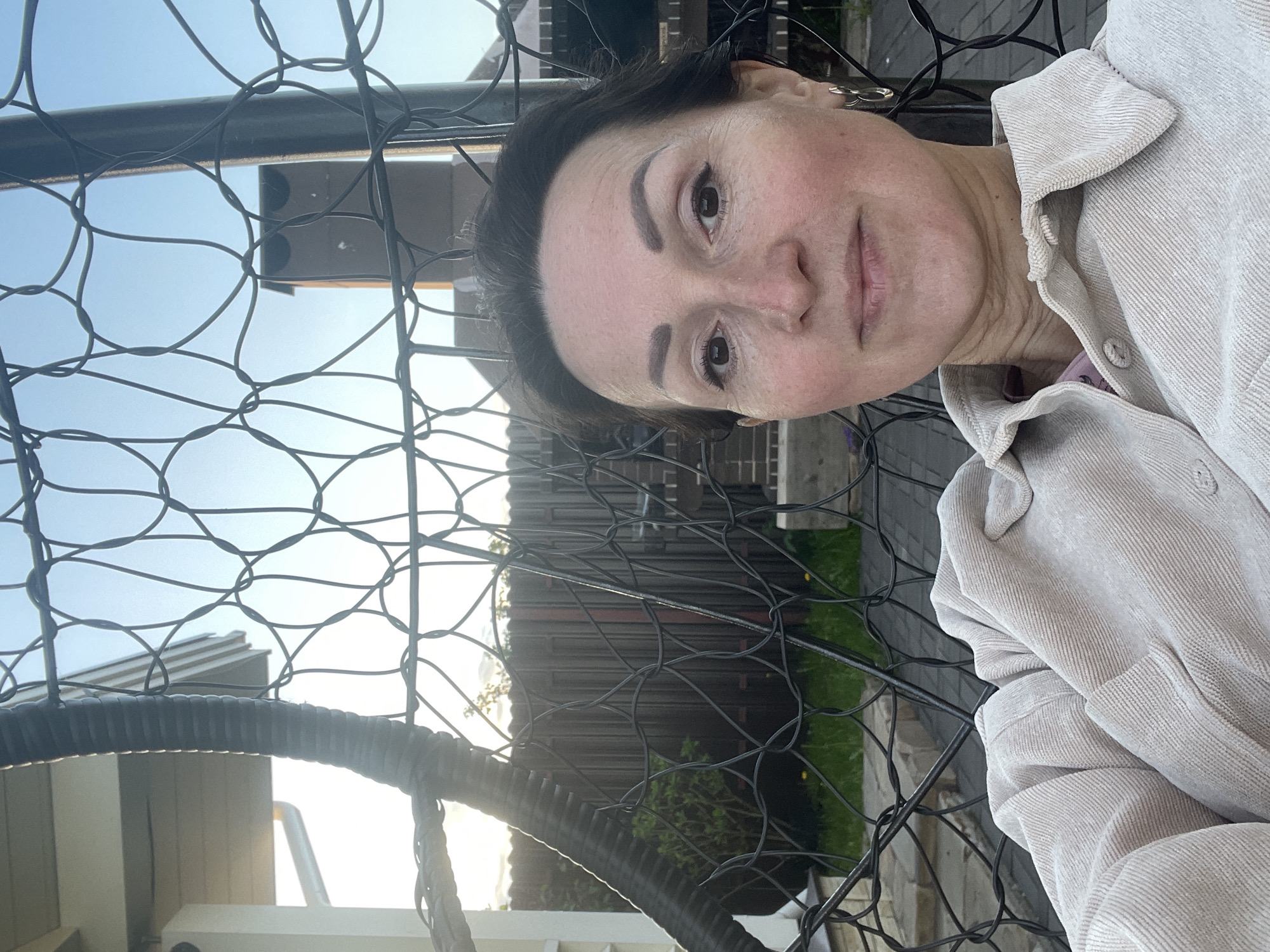 A woman is photographed at an outdoor location in Solov'evka, Russia. The subject is a middle-aged woman wearing a light-colored corduroy jacket. A black wire mesh fence or enclosure partially obscures the view. Behind the fence, there is a wooden structure, possibly a house, and some greenery, suggesting a garden or backyard setting. The lighting indicates it might be late afternoon or early evening.