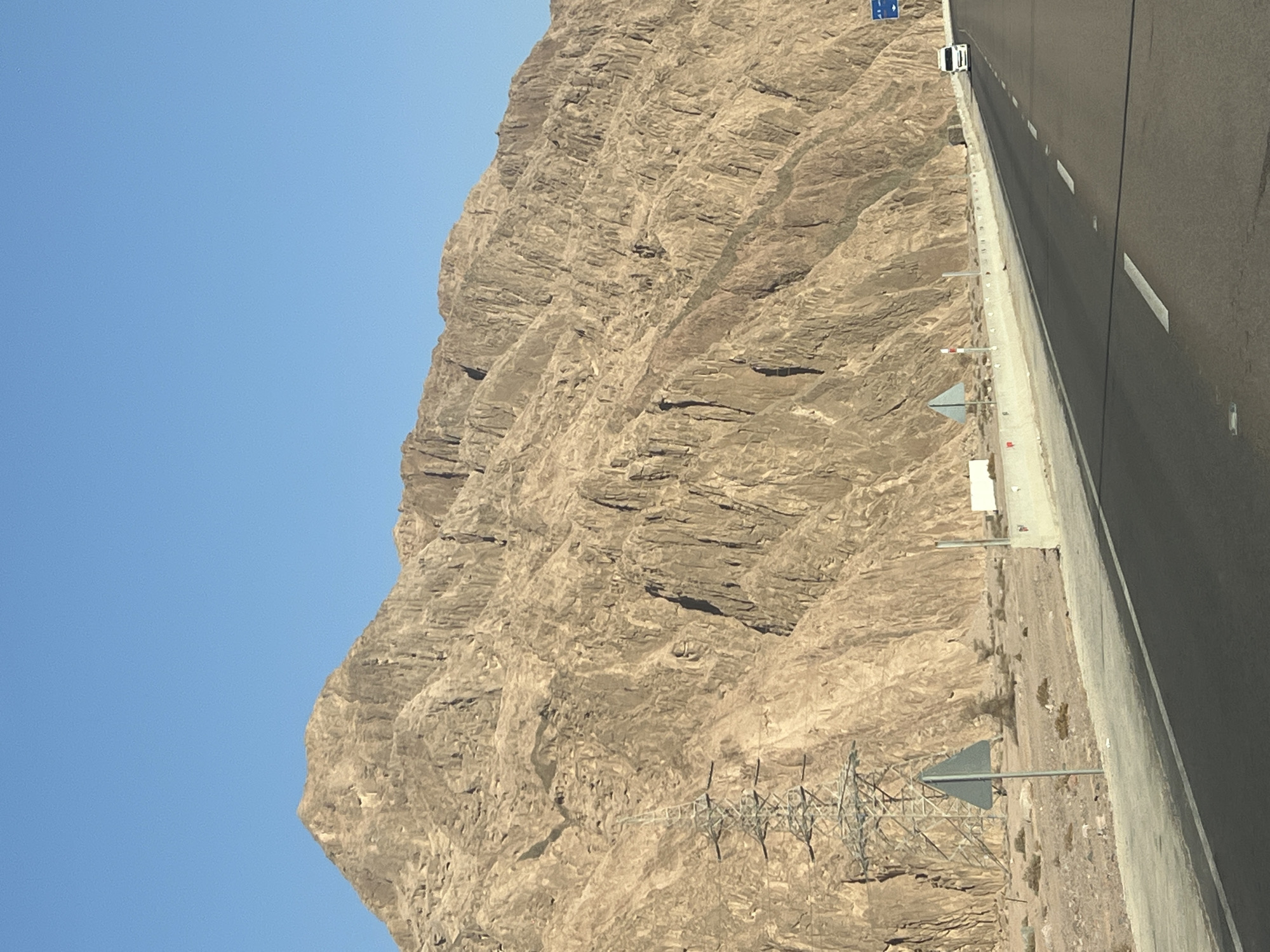 A clear, sunny day in Dahab, Egypt, features a vast, arid mountain range dominating the background. The mountains are rugged and textured, with deep fissures and a tawny, rocky surface. In the mid-ground, a modern highway runs parallel to the mountains, with a white concrete barrier separating the road from the desert landscape. To the left of the highway, a large, skeletal metal electricity pylon stands against the mountainside, with warning signs in front of it. A few sparse desert bushes dot the sandy terrain. On the highway, a white van is visible in the distance, traveling in the same direction as the viewer. The sky is a vivid, cloudless blue.