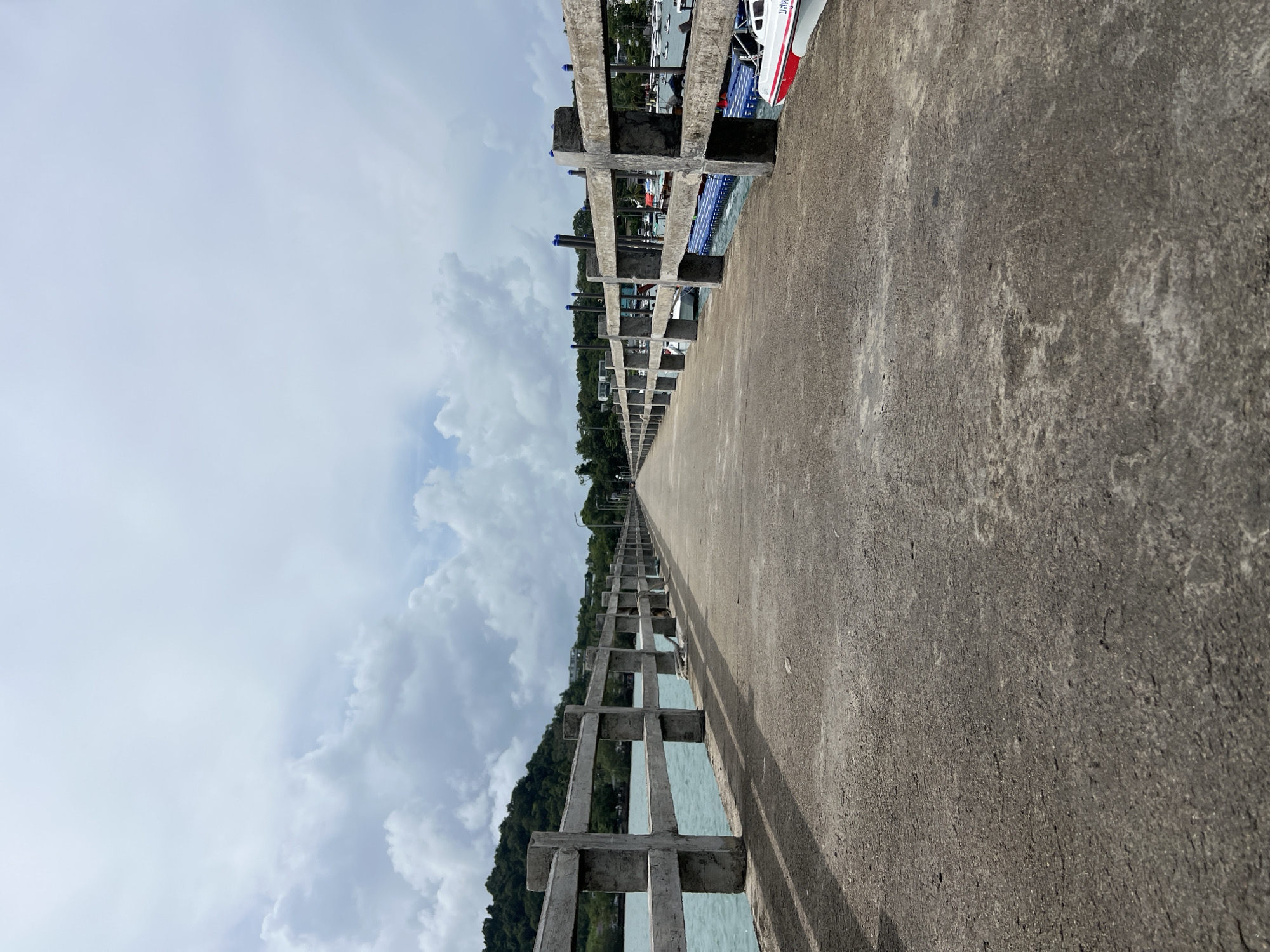 A long, concrete pier extends into the distance under a cloudy sky in Pa Khlok, Thailand. The pier has a concrete railing on both sides, supported by posts. To the left of the pier, the turquoise water is visible, with a lush, green, tree-covered hillside in the background. To the right, several boats are docked, including a white and red speed boat in the foreground. The pier appears to be a public walkway or a docking area for boats.