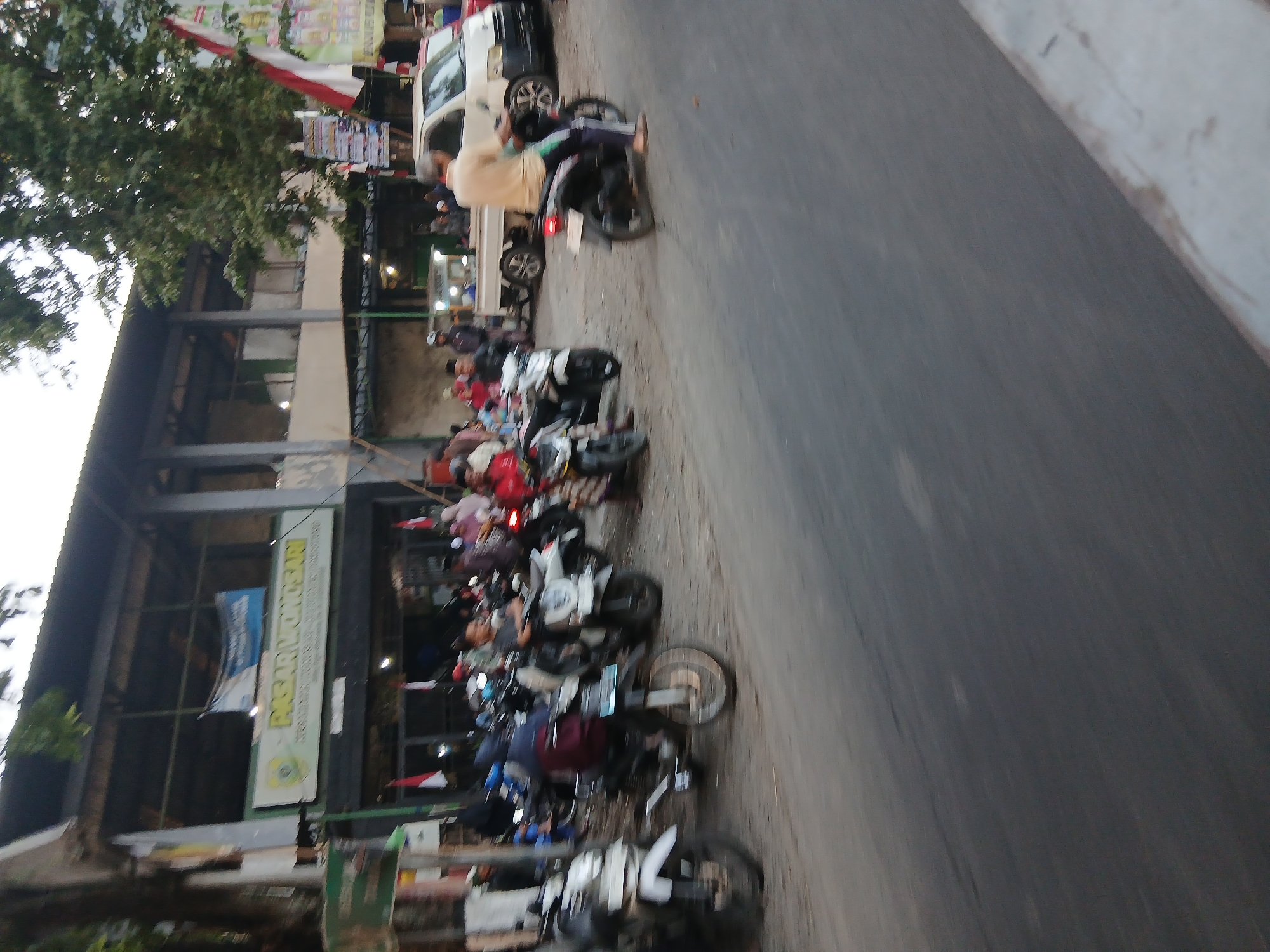 The image shows a street scene in Wonosari, Indonesia. A building with a sign that reads "PASAR WONOSARI" (Wonosari Market) is visible. Several people are present, many of them on motorcycles. Some are parked, while others appear to be in motion. A white van is also parked on the street. The environment appears to be an outdoor market area with a road in the foreground. The image is taken from a slightly elevated perspective, looking across the street towards the market. There are trees and flags visible in the background.