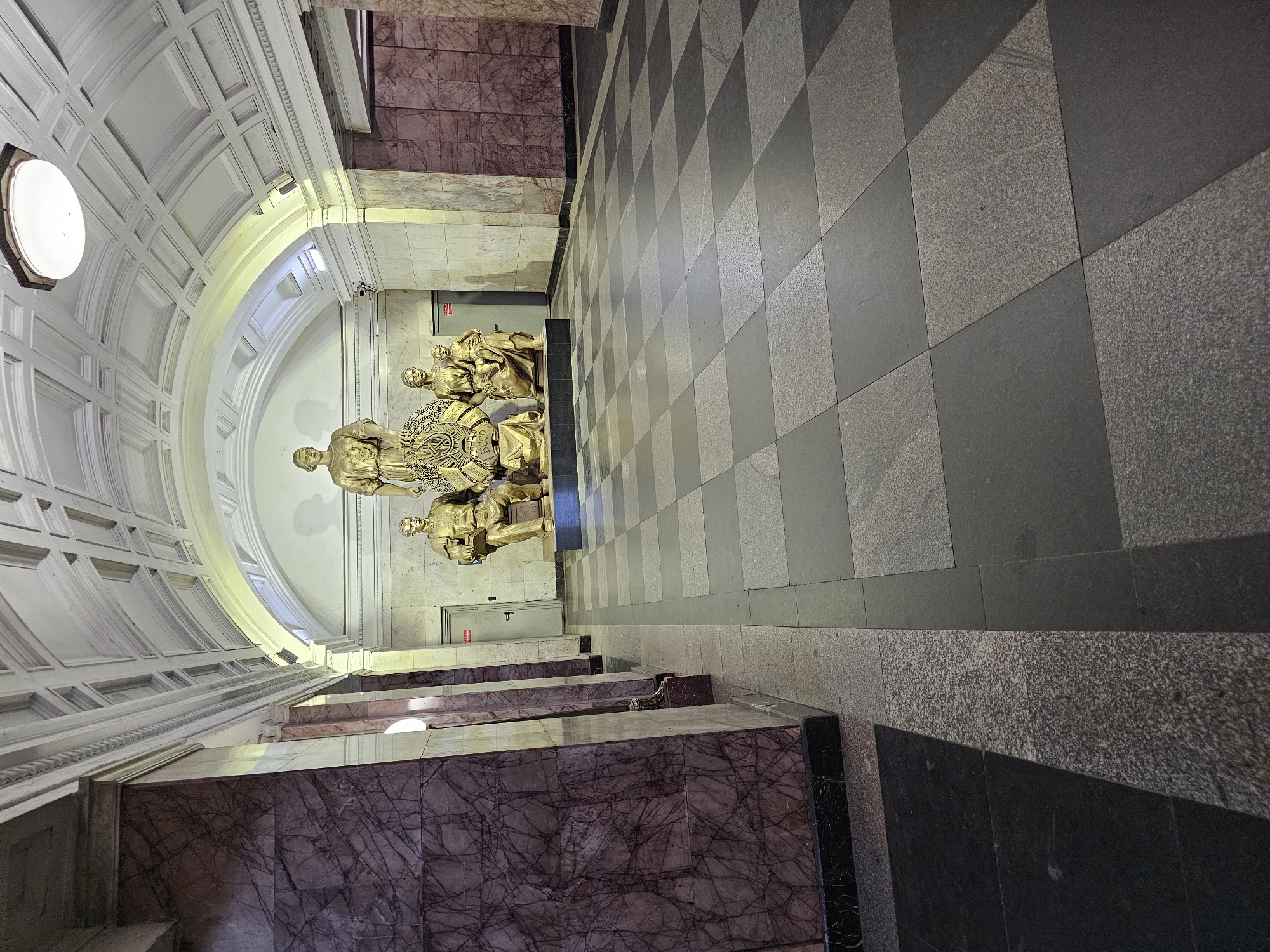 The image depicts the Ploshchad Revolyutsii station in Moscow, Russia. Visible are golden statues depicting Soviet people. The platform is marked by a checkered pattern on the floor. Marble-clad staircases with veined patterns are also visible. The architecture features arched ceilings.