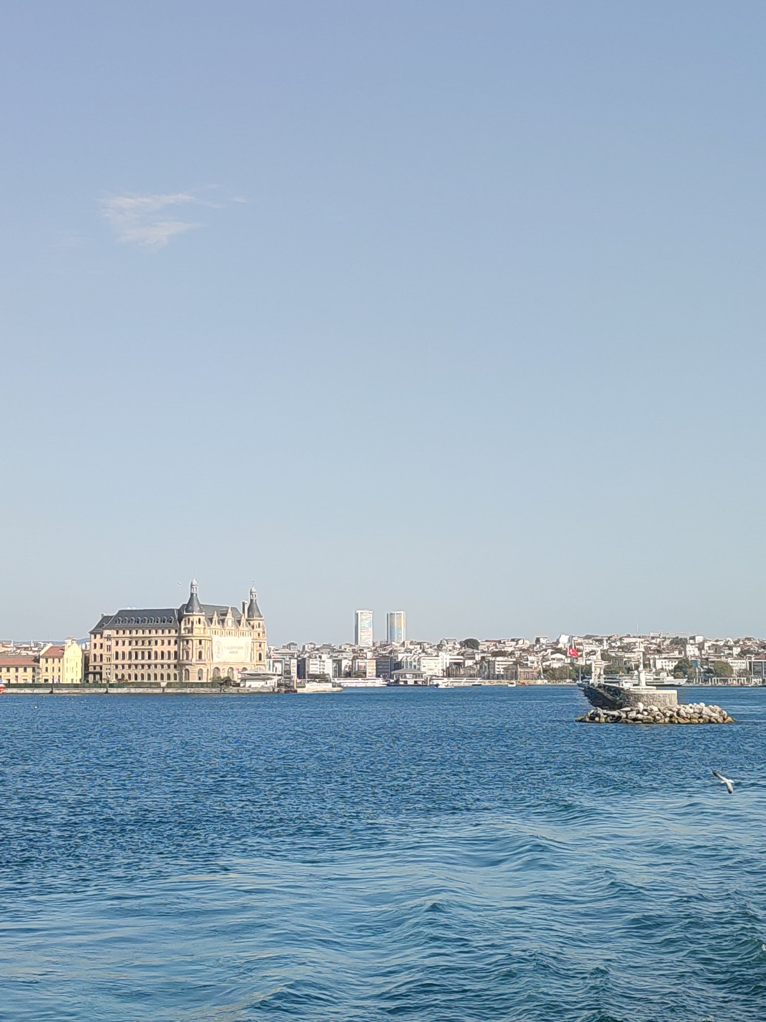 The image captures a waterfront view of the Haydarpaşa Train Station in Istanbul, Turkey, from a boat on the water. The historic train station, a prominent landmark, is situated on the left side of the frame, characterized by its distinctive architecture with multiple stories, arched windows, and twin towers. A cityscape with numerous buildings, including some modern high-rises, stretches across the horizon behind the station. The foreground is dominated by the blue water of the Bosphorus, with gentle waves creating a rippled texture. On the right side of the image, a small rocky pier juts out into the water, topped with a monument or sculpture. A Turkish flag is visible on the pier. A bird is seen flying in the middle-right of the frame. The sky is clear and light blue with a few wispy clouds.