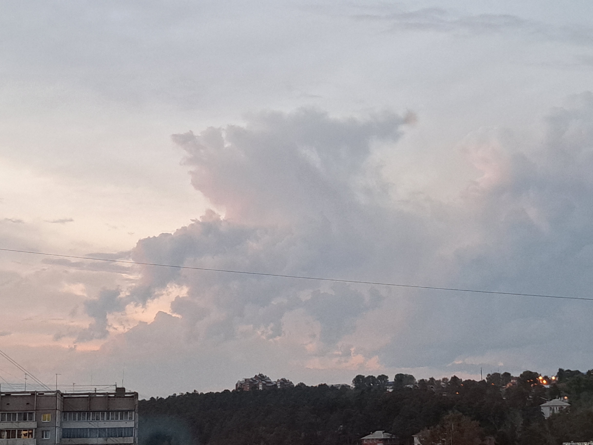 The image captures a panoramic view of the sky and cityscape in Irkutsk, Russia. The sky is filled with soft, diffused light, characteristic of twilight, with clouds displaying a mix of pale gray and hints of pink and peach hues. A thin, dark power line stretches horizontally across the upper portion of the frame, intersecting the sky.
In the lower part of the image, a multi-story residential building is visible on the left. It appears to be constructed from concrete panels, with numerous windows arranged in rows. The building exhibits a somewhat utilitarian architectural style. To the right of the building, a dense, dark green forest covers a hillside. Scattered amongst the trees, the lights of houses and other structures can be seen, suggesting a residential area nestled on the incline. The overall impression is one of a quiet evening scene in a Russian city, blending urban development with natural landscapes.