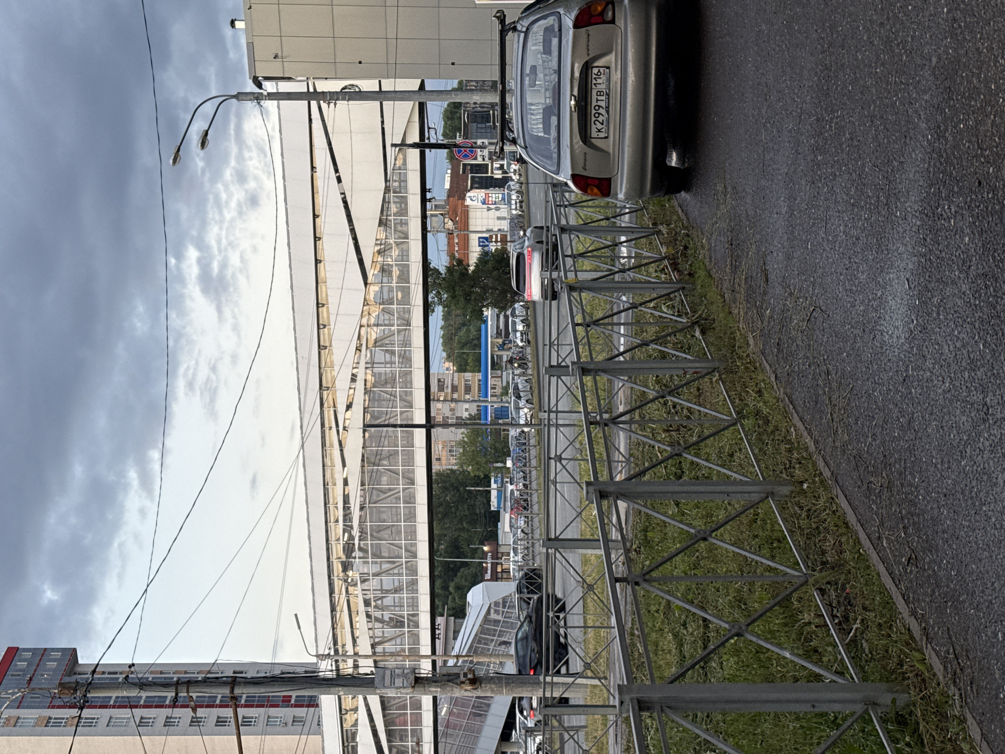 A medium shot captures a street scene in Kazan, Russia, under a cloudy sky. In the foreground, a silver Chevrolet car with license plate K299TB 116 is parked on a wet asphalt road, facing away from the viewer. A metal fence runs along the side of the road, separating it from a grassy area. In the midground, a modern pedestrian bridge with a glass enclosure spans across the road, connecting two sides of the city. Vehicles are visible on the road, some in motion. Behind the bridge, several buildings of varying heights and architectural styles can be seen, including a tall, modern residential building on the left. The overall impression is one of urban infrastructure and daily city life.