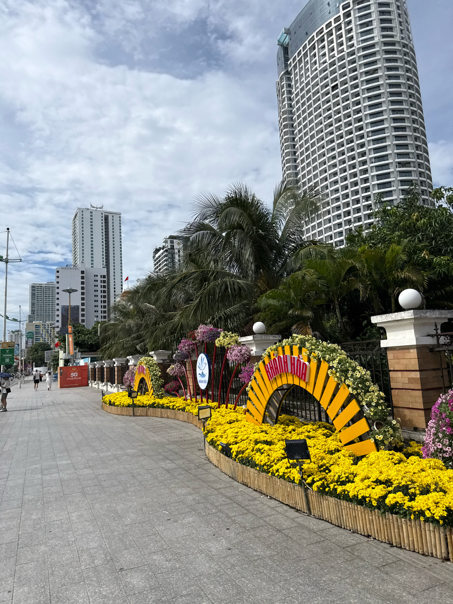 The image depicts a street scene in Nha Trang, Vietnam, likely a tourist area, given the presence of palm trees and tropical vegetation alongside modern high-rise buildings. In the foreground, a paved walkway curves along a decorative floral arrangement. This arrangement features a semicircular yellow structure adorned with yellow and green flowers, and text in red, appearing to spell "KHANH HOA". Behind the floral display, several tall, modern buildings dominate the skyline, with one particularly large, curved skyscraper with many windows. Palm trees and other greenery are interspersed between the buildings and in front of them. A few people are visible walking on the pavement in the background, some holding umbrellas, suggesting it might be overcast or a light rain. There are also streetlights and signage, including a "5G mobifone" advertisement. The sky is cloudy.