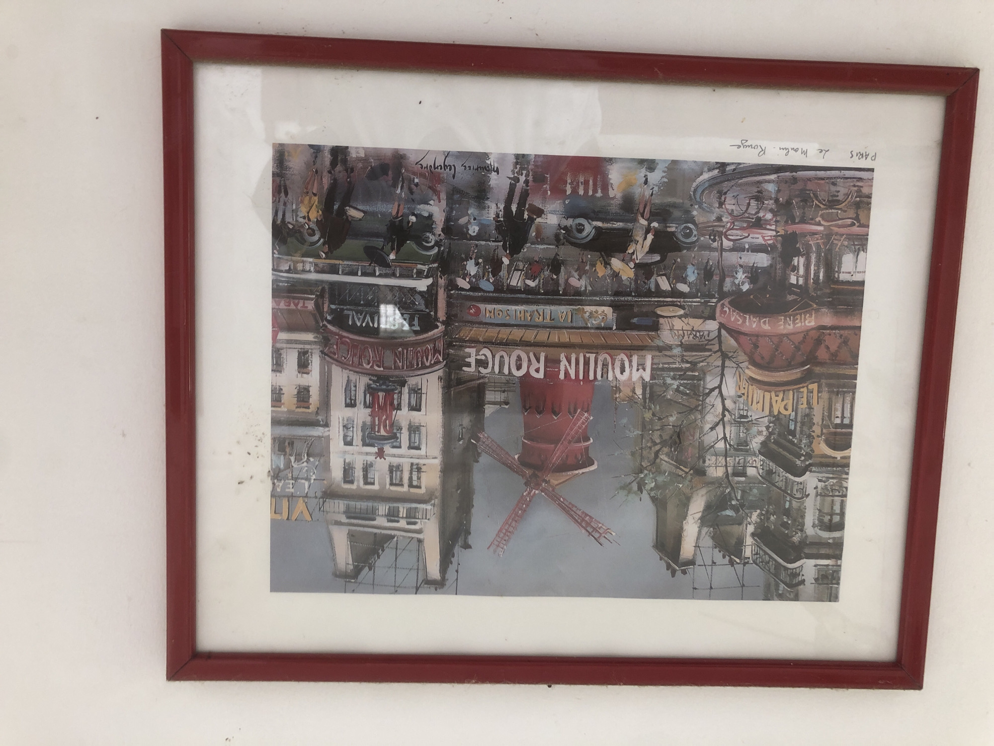 The image is a framed painting of the Moulin Rouge in Paris. The painting depicts a rainy street scene with the iconic red windmill visible in the background. People with umbrellas are walking on the wet street, and there are cars and a bus. Buildings with advertisements for "Moulin Rouge" and "Festival" line the street. In the foreground, the text "PARIS Le Moulin Rouge" is handwritten below the painting. The frame of the painting is red. There is no information in the image that suggests any connection to La Tagua, Venezuela.