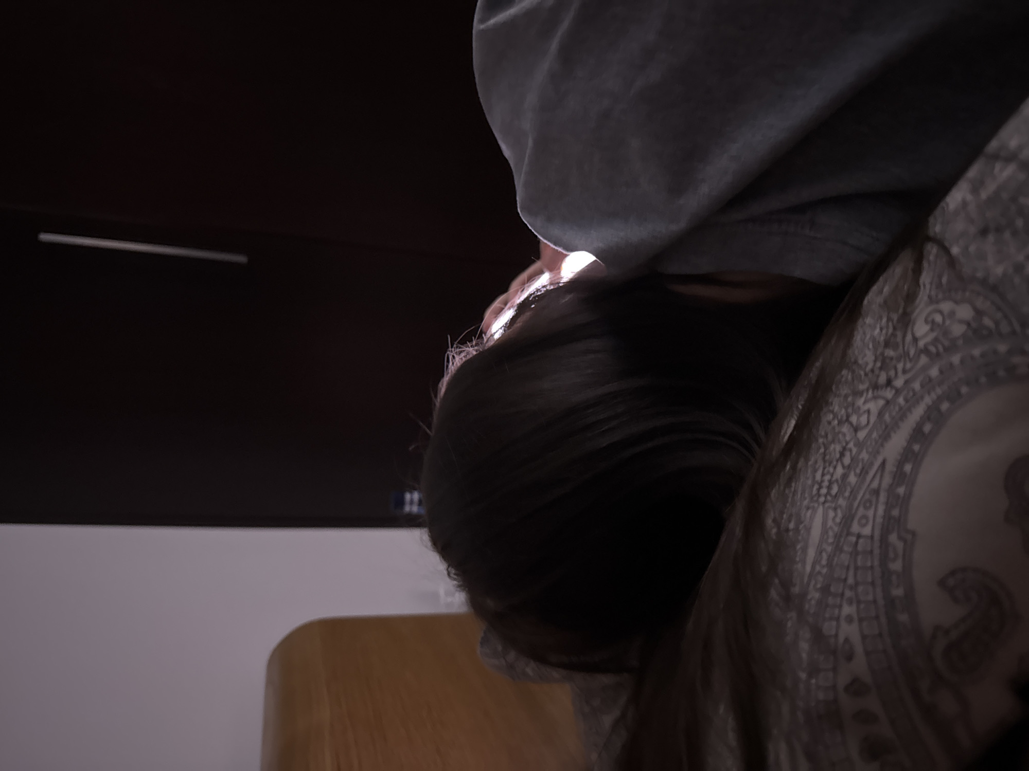A woman is lying down in a bed. The woman is wearing a gray sweatshirt. The bed has a gray paisley patterned pillowcase. A dark brown headboard is behind the woman. The location of the image is unknown, but the request mentions the city of Warsaw, Poland. The scene is dark, with a light source illuminating the woman's face.