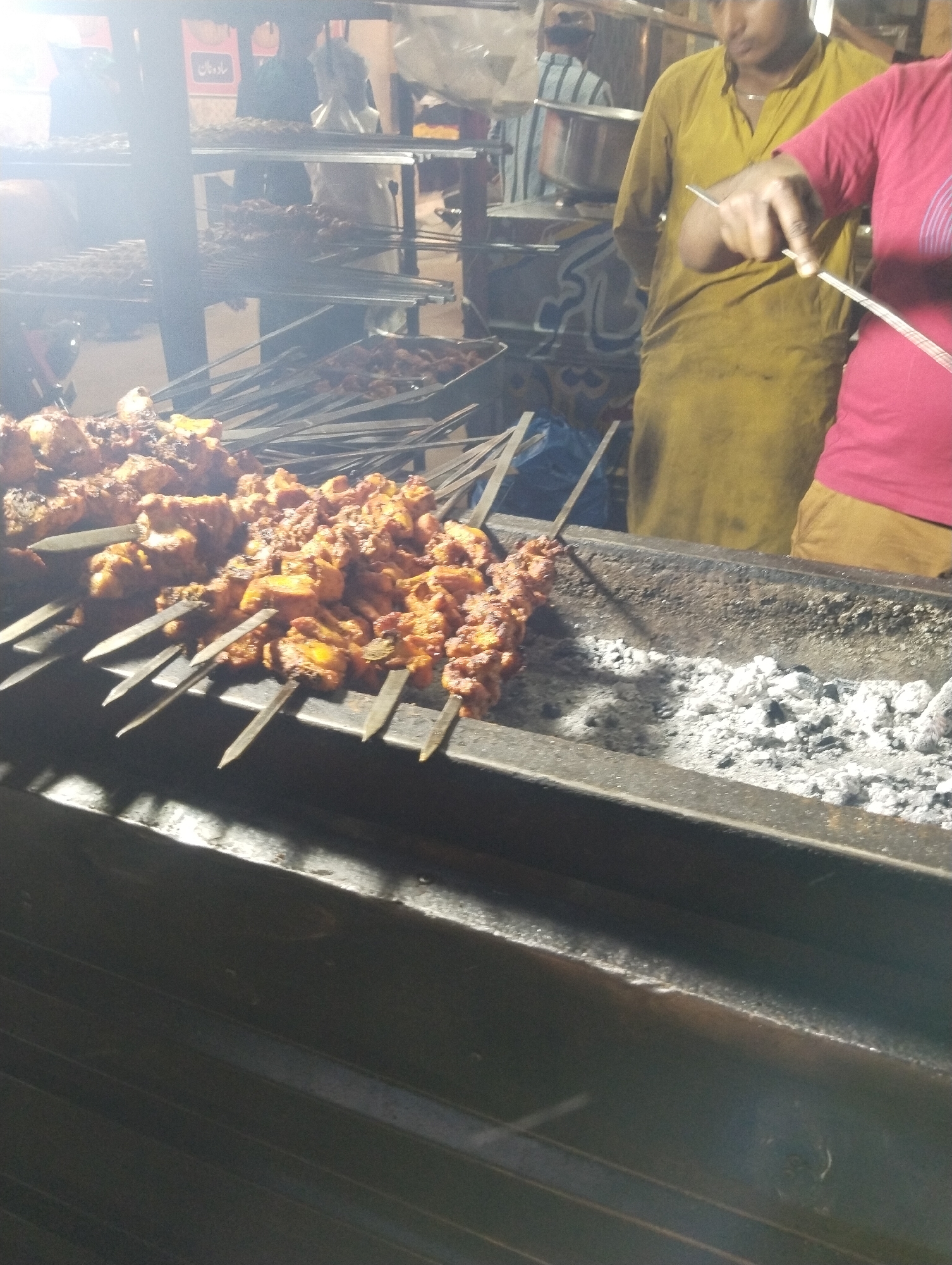 The image depicts a vibrant nighttime scene at a food stall, likely in Heir, Pakistan, specializing in grilled meats. Numerous skewers, laden with marinated chicken or meat pieces (kebabs or tikka), are actively cooking over a long, rectangular charcoal grill. White and grey ash from burning coals is visible within the grill, and smoke or steam rises from the cooking food.
Two primary figures are visible. On the right, a person wearing a red t-shirt holds a long, thin tool, seemingly managing the skewers. Behind them, another person dressed in a mustard yellow traditional garment (likely a shalwar kameez) stands, observing the grilling process. In the background, additional skewers are arranged on racks, and other individuals are faintly visible, including one in a striped shirt.
The strong artificial lighting illuminates the cooking area, creating bright highlights and a slight lens flare in the upper left, indicating it is dark outside. The setting suggests a bustling street food environment or an outdoor eatery. A red sign with white Arabic script is visible in the upper background, displaying the text "ساوہ نان" (possibly "Sawoh Naan" or "Green Bread"), which points to a naan or bread-making component of the establishment. The scene captures the essence of traditional South Asian grilled cuisine.