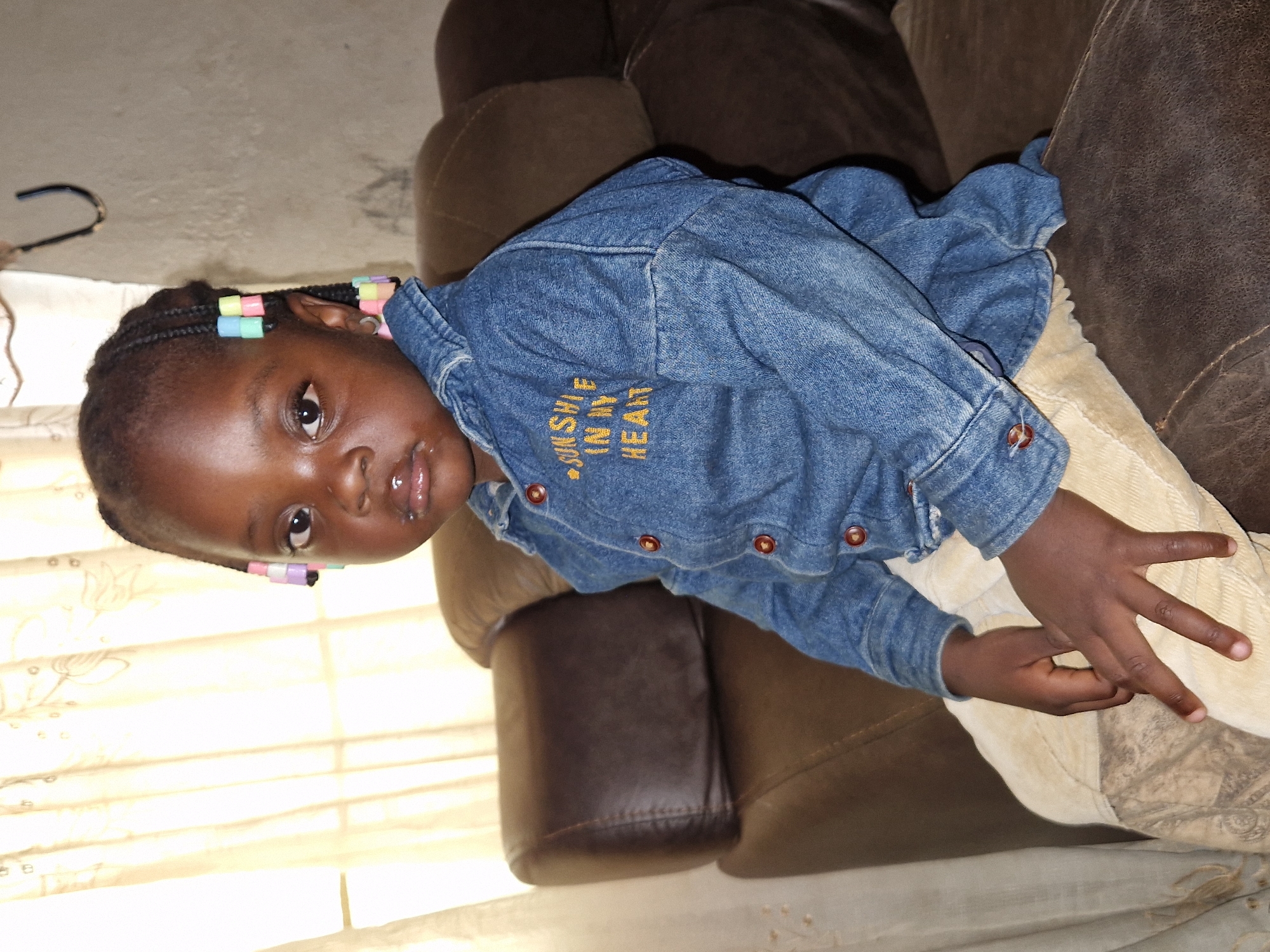 A young Black child is seated on a brown leather couch, facing forward and slightly to the right. The child has braided hair adorned with colorful beads. They are wearing a denim shirt with yellow embroidery that reads "SUNSHINE IN MY HEART" and light-colored pants. The background includes a cream-colored curtain and a light-colored wall with a black hook. The image appears to be taken indoors in a residential setting. The lighting suggests a diffused natural light source, possibly from a window. The scene is likely located in Jos, Nigeria, based on the provided context.