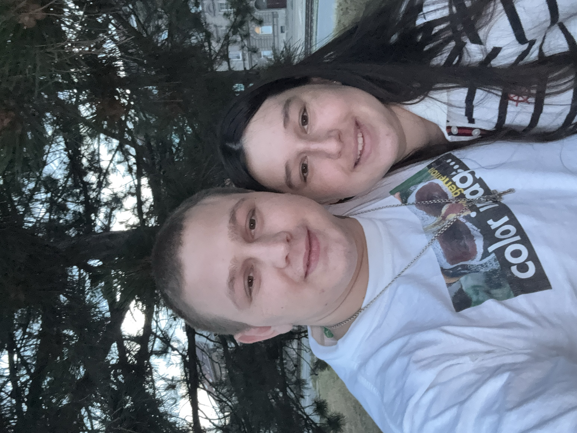 A selfie taken outdoors features a young couple in the foreground, positioned slightly off-center to the left. The man, on the left, has light skin, short brown hair, and is wearing a white t-shirt with a graphic design and a silver chain. The woman, on the right, has light skin, long dark hair, and is wearing a black and white striped collared shirt. They are both smiling and appear to be embracing or standing close together.
Behind them, a dark evergreen tree with dense branches fills the upper portion of the frame, with a glimpse of the sky visible through the foliage. In the background, a multi-story building with visible windows and architectural details can be seen. The overall setting suggests an outdoor urban or park environment, possibly in Pilsen, Czechia, given the architectural style of the building. The lighting indicates it is either early morning or late evening.