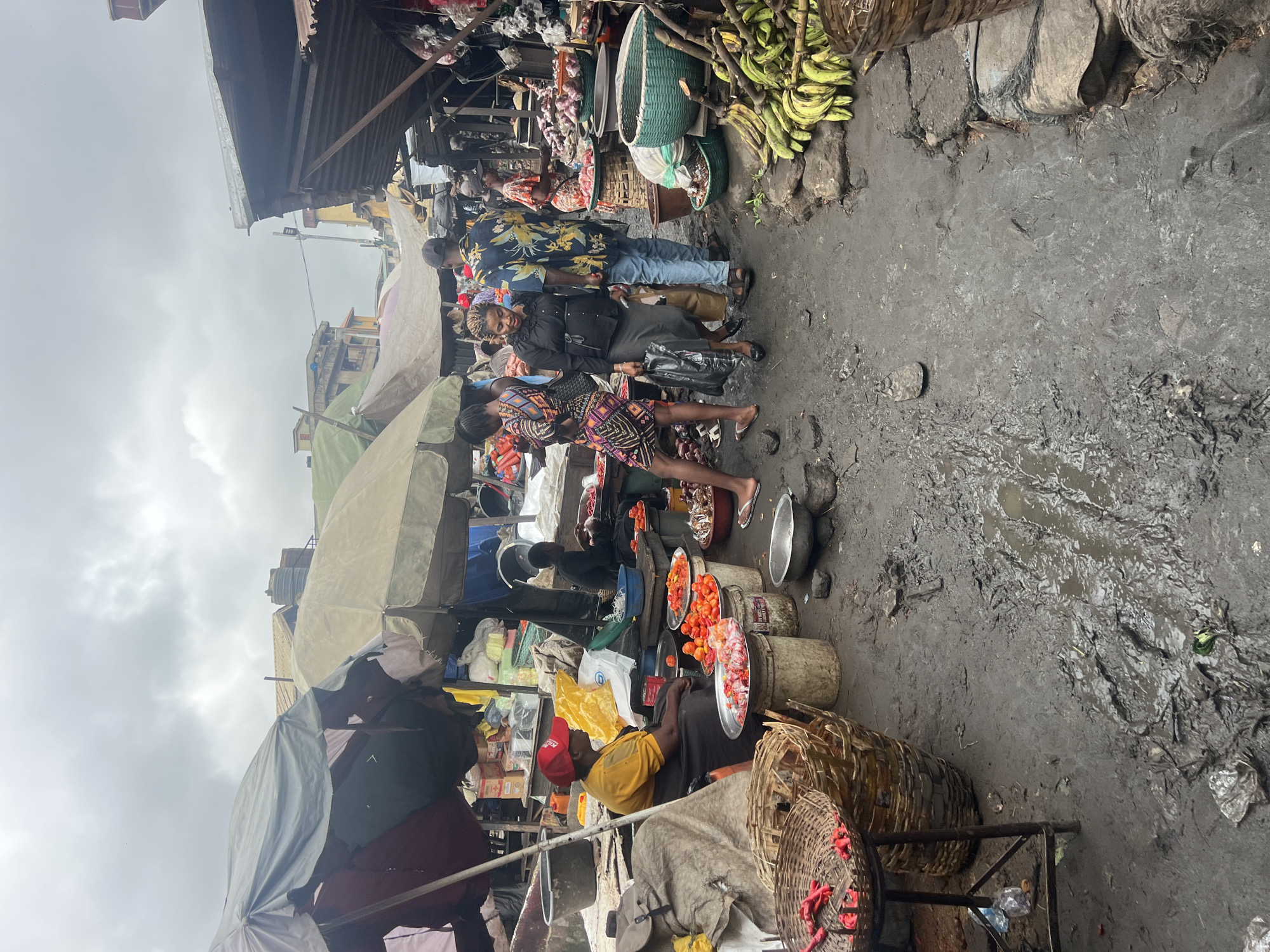 The image depicts a bustling outdoor market, possibly in Nkpor, Nigeria. The ground is muddy and uneven, with puddles of water visible. Numerous market stalls are set up, covered by tarpaulins and umbrellas to provide shade.
Several people are present, engaged in various activities:
* **Merchants:** A woman in a yellow shirt and red cap sits near a display of tomatoes and chili peppers, appearing to be a vendor. Another person, dressed in black, is seated behind a stall filled with red peppers and other produce.
* **Shoppers/Passersby:** A group of individuals are seen walking through the market. One woman in a black dress and grey skirt is carrying a black plastic bag. A man in a floral shirt and blue jeans walks beside her. Another woman in a patterned dress is also present.
The market is filled with a variety of goods:
* **Produce:** Piles of tomatoes, red chili peppers, onions, and bunches of green bananas are prominently displayed.
* **Baskets and Containers:** Woven baskets are used to hold produce, and various metal bowls and plastic containers are scattered throughout the scene.
* **Shelter:** Tarpaulins and umbrellas provide cover for the stalls and merchandise.
* **Environment:** The market appears to be in a somewhat disorganized state, with debris and muddy patches on the ground. Buildings are visible in the background, suggesting an urban or semi-urban setting. The sky is overcast.