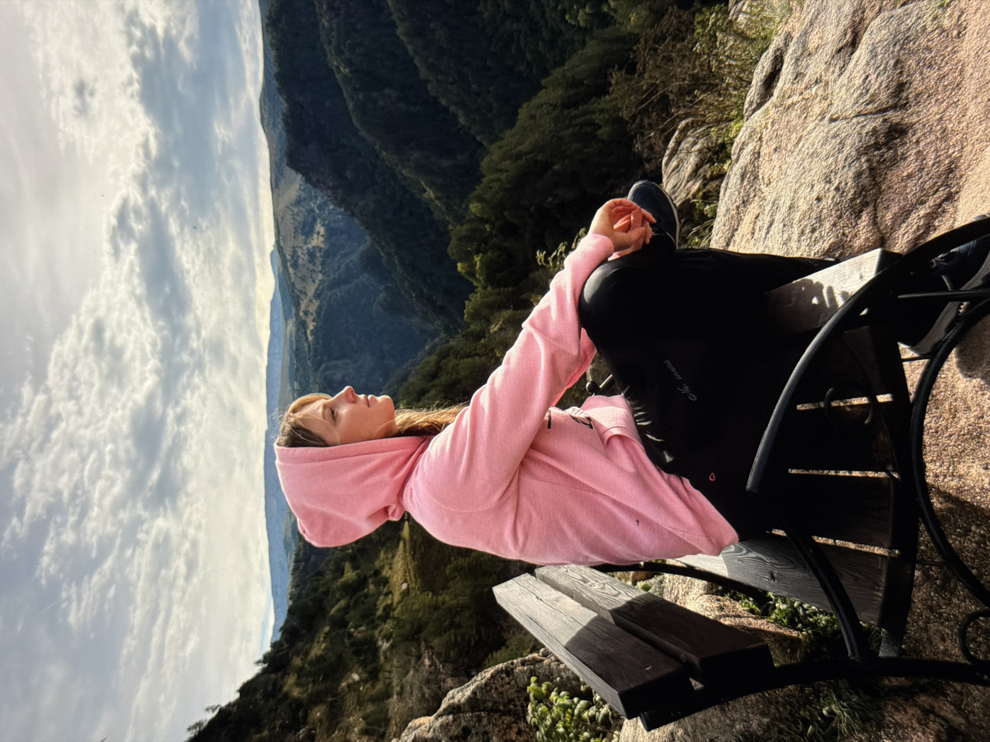 A young woman wearing a pink hooded sweatshirt and black pants sits on a wooden bench on a rocky overlook, gazing at a vast, tree-covered mountain valley under a cloudy sky. The bench is made of dark wood and wrought iron. The landscape features rugged terrain with dense green trees and distant, hazy mountains. The lighting suggests it is daytime, possibly late afternoon due to the long shadows. There are no visible landmarks to indicate this location is within the city of Krasnodar, Russia.