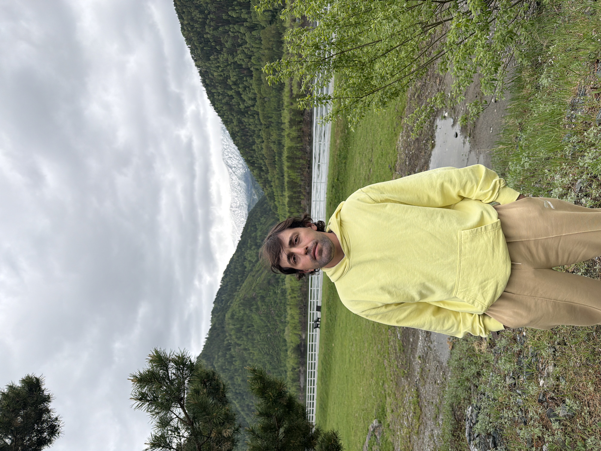 A person stands on a grassy area with a mountain range in the background. The person is wearing a yellow hoodie and khaki pants. A white fence is visible behind the person, with a body of water and trees. The sky is cloudy. It is not possible to determine if the image was taken in Cheremushki, Russia.