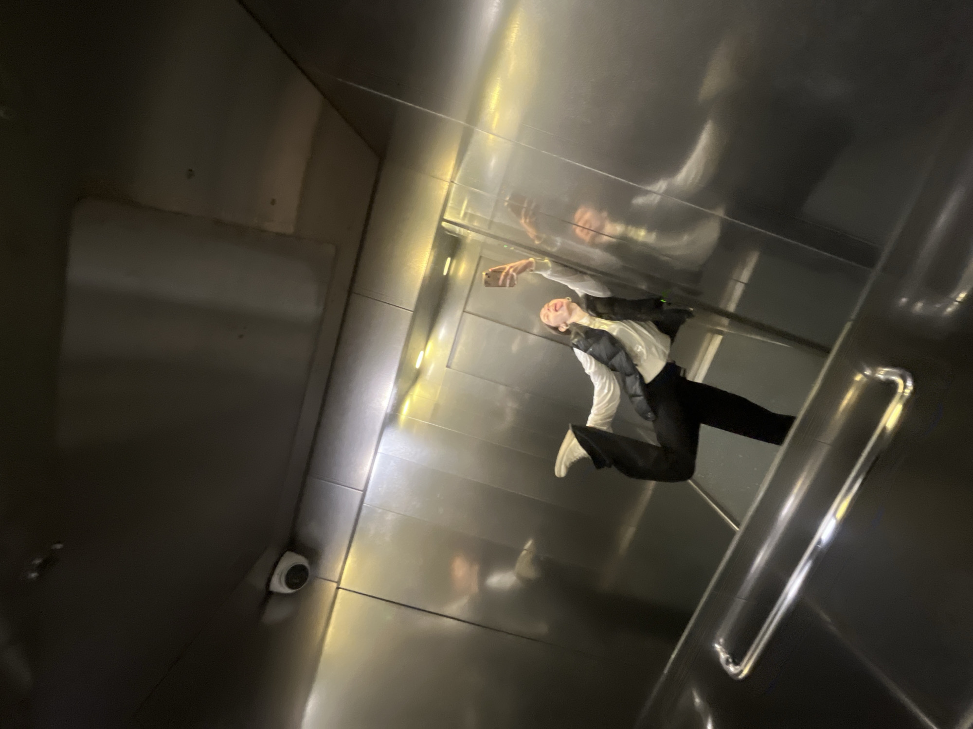 A person wearing a white shirt, dark vest, and dark pants is standing in an elevator, holding a phone up and taking a selfie. The elevator has metal walls and an integrated security camera. The person is reflected in the metal walls. The location is Saint Petersburg, Russia.