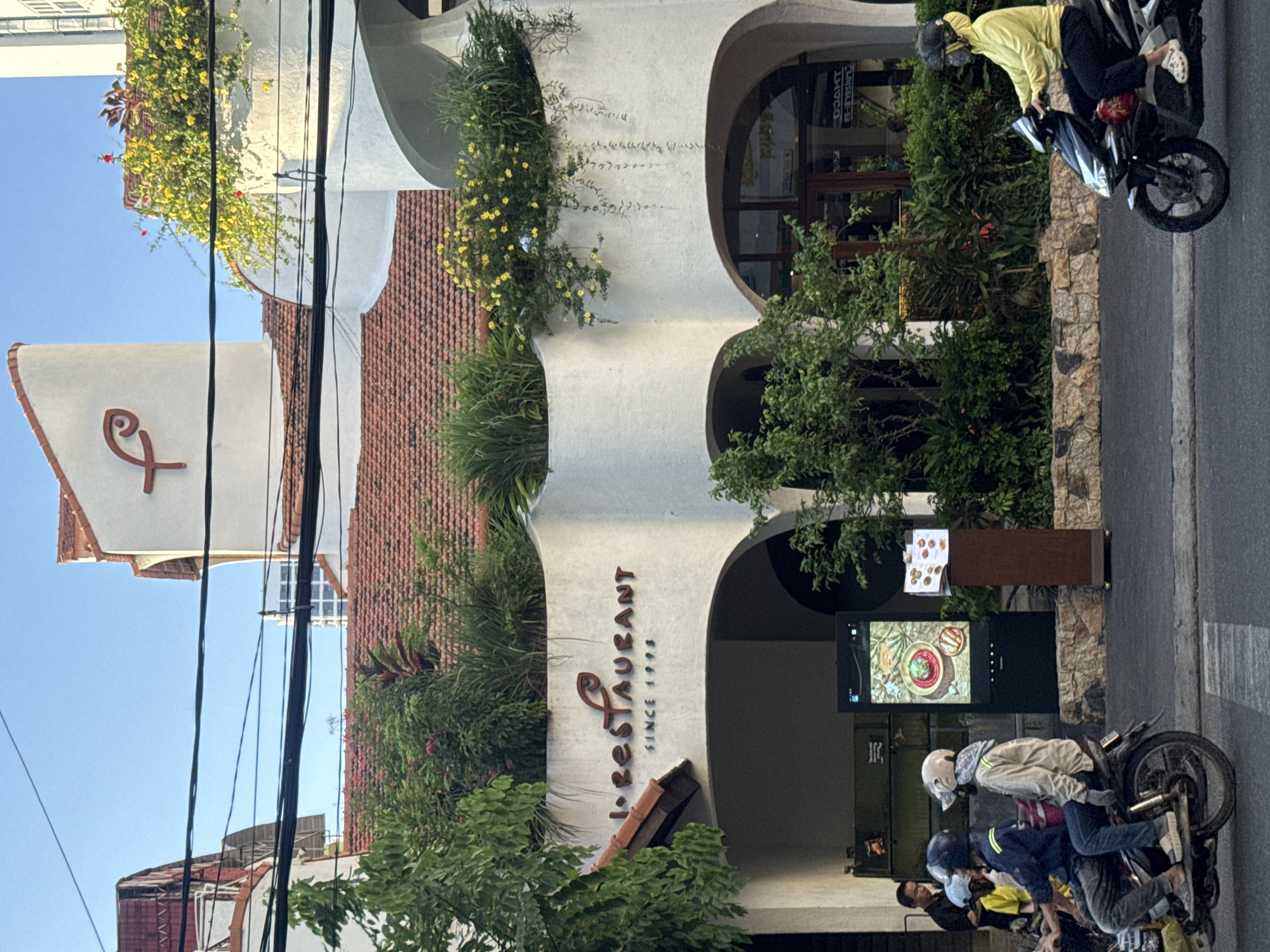 The image shows a street scene in Nha Trang Ward, Vietnam, in front of a white building with a curved facade and a red tile roof. The building has a large sign that reads "L. RESTAURANT SINCE 1998" with a stylized fish logo above the text. Greenery, including bushes and flowering vines, is visible on ledges and around the building.
In the foreground, several people are on motorcycles. Two individuals are riding on a motorcycle on the left, and another person is riding a motorcycle on the right. The person on the right is wearing a bright yellow hooded jacket and black pants.
A digital display screen is positioned on the sidewalk in front of the restaurant, showing a picture of a dish. Next to the display is a small wooden stand with what appears to be a menu.
The street is paved, and power lines are visible across the top of the image. The sky is clear and blue, suggesting it is daytime. The architecture of the building, with its curved lines and the prominent signage, suggests it is a restaurant or commercial establishment.