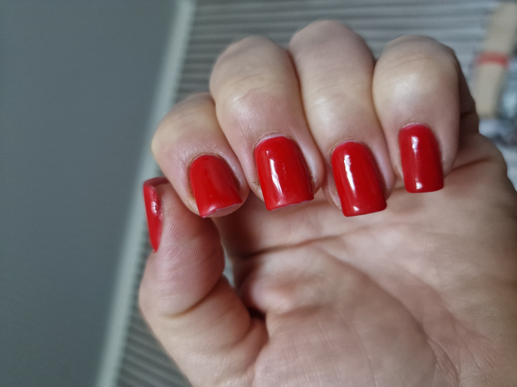 A close-up, low-angle shot captures a left hand with bright red nail polish. The hand is positioned against a blurred background of grey and white vertical stripes, suggesting a wall or textured surface. The lighting is soft, highlighting the glossy finish of the nails and the natural texture of the skin. The image appears to have been taken indoors, possibly in a residential setting. There is no visible indication of the location being Kursavka, Russia, or any other specific geographical area.