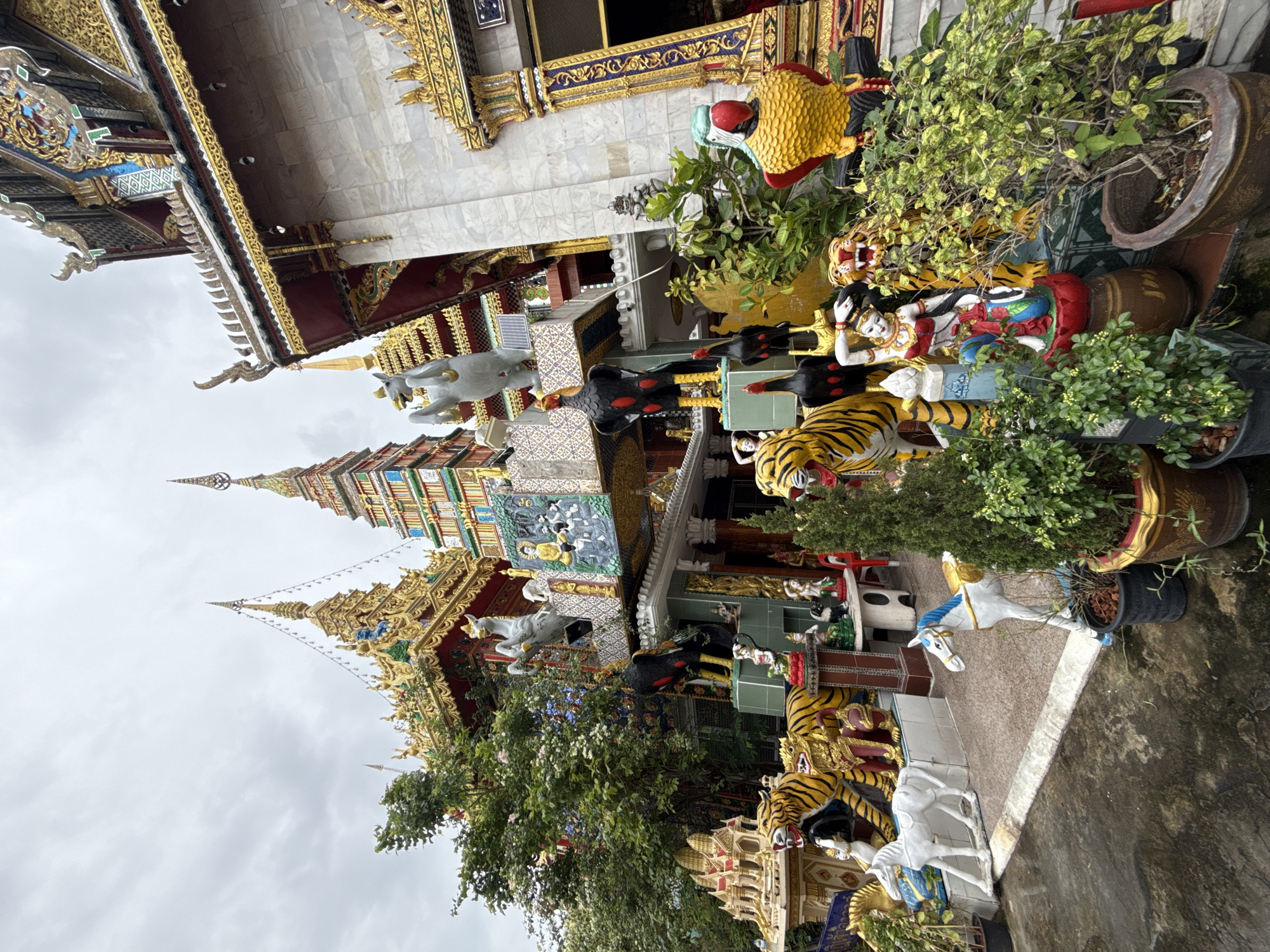 The image depicts the exterior of a temple in Bangkok, Thailand, characterized by its ornate architecture and numerous decorative statues. The temple features multi-tiered roofs with golden spires and intricate detailing, typical of Thai Buddhist temple design. Prominently displayed are various vibrant statues, including tigers, roosters, horses, and human figures, which adorn the temple grounds and facade. Some of these statues are life-sized, adding to the visual spectacle. The background shows a cloudy sky, suggesting overcast weather. The scene is an outdoor environment with paved ground and some greenery, including potted plants. The overall impression is one of a richly decorated and symbolically significant religious site.